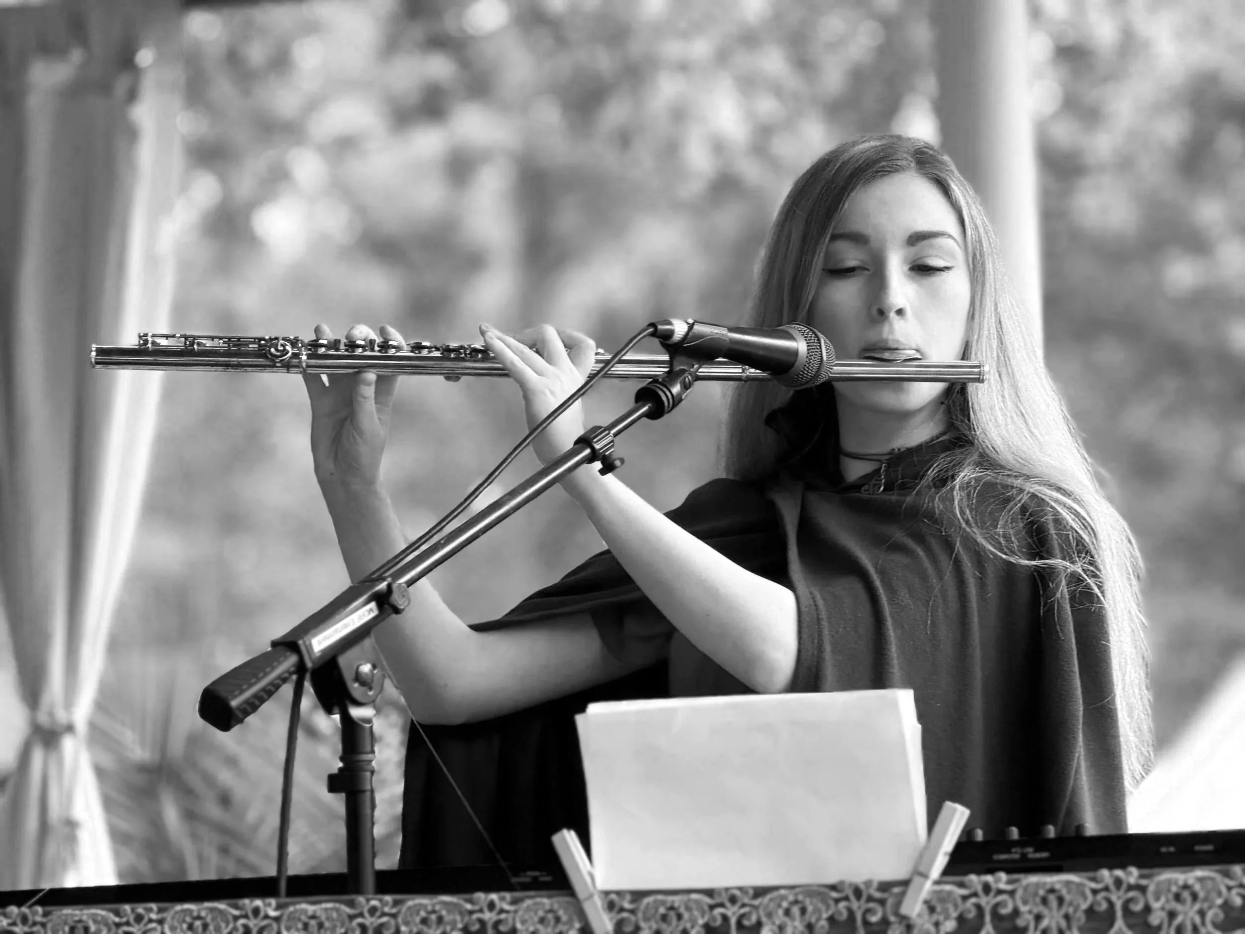 A young woman playing a flute at a microphone, with music sheets in front of her, in an outdoor setting.