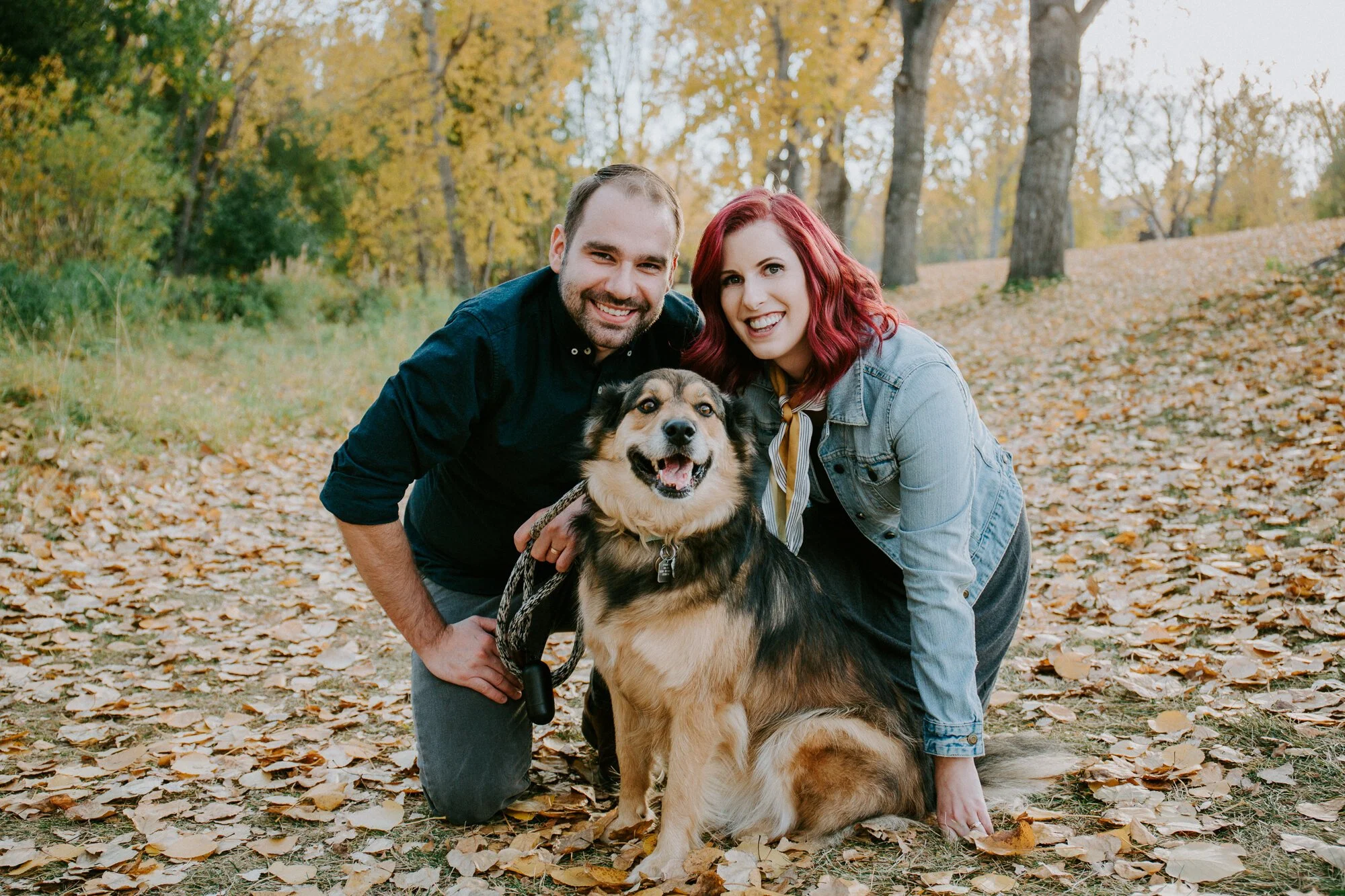 A couple kneeling beside a dog on a leaf-covered path in an autumn forest.