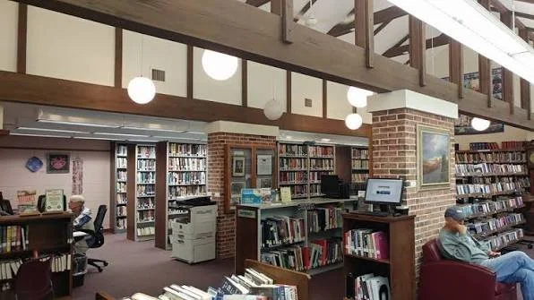 Interior of a public library with bookshelves, a couple of people reading, and a computer station.