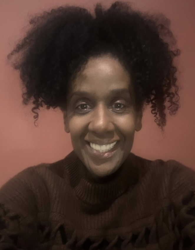 A woman with dark, curly hair smiling in front of a pink wall.