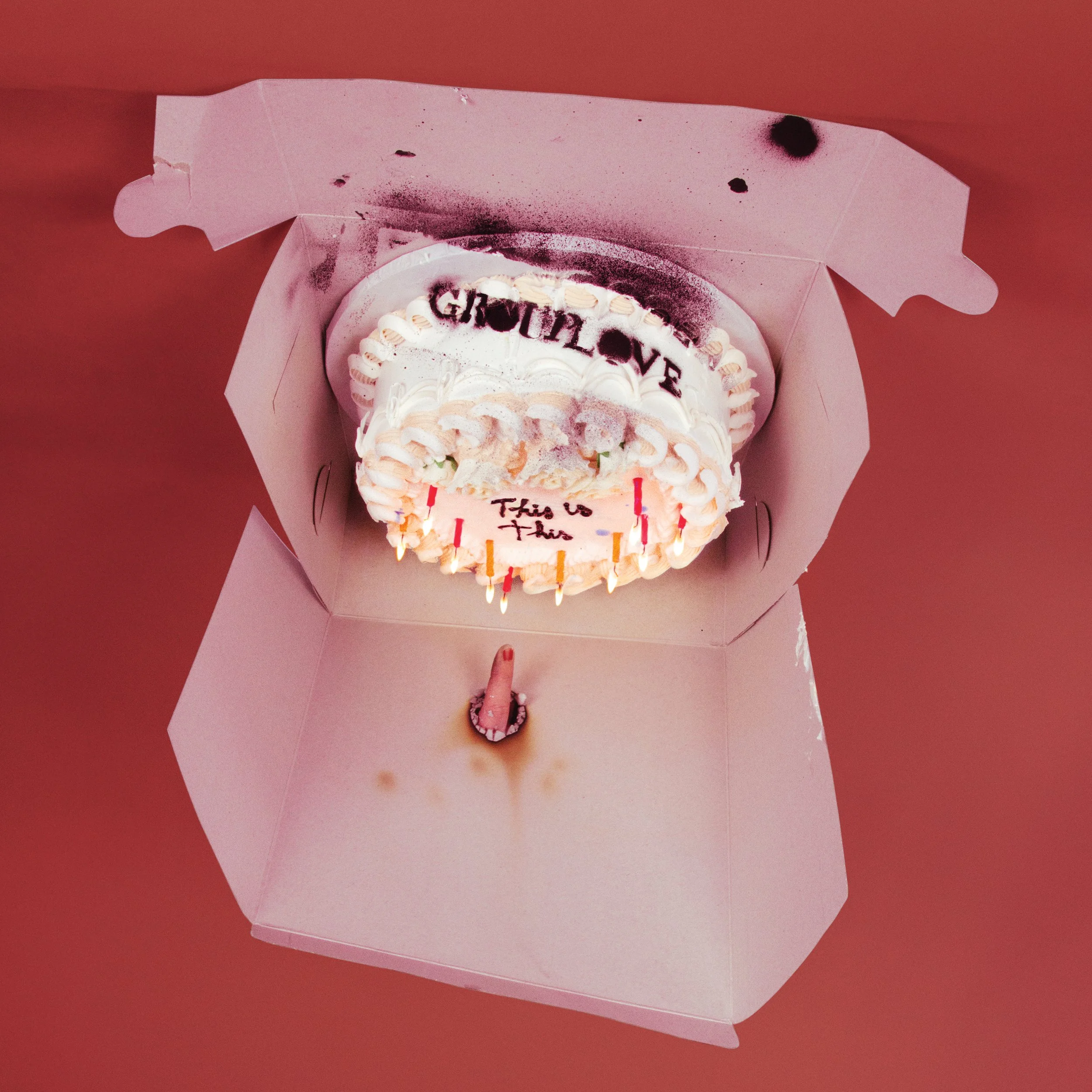 A birthday cake with white frosting and candles inside a pink cake box, with red and black text that reads 'GROUPLOVE' and 'This is this.'