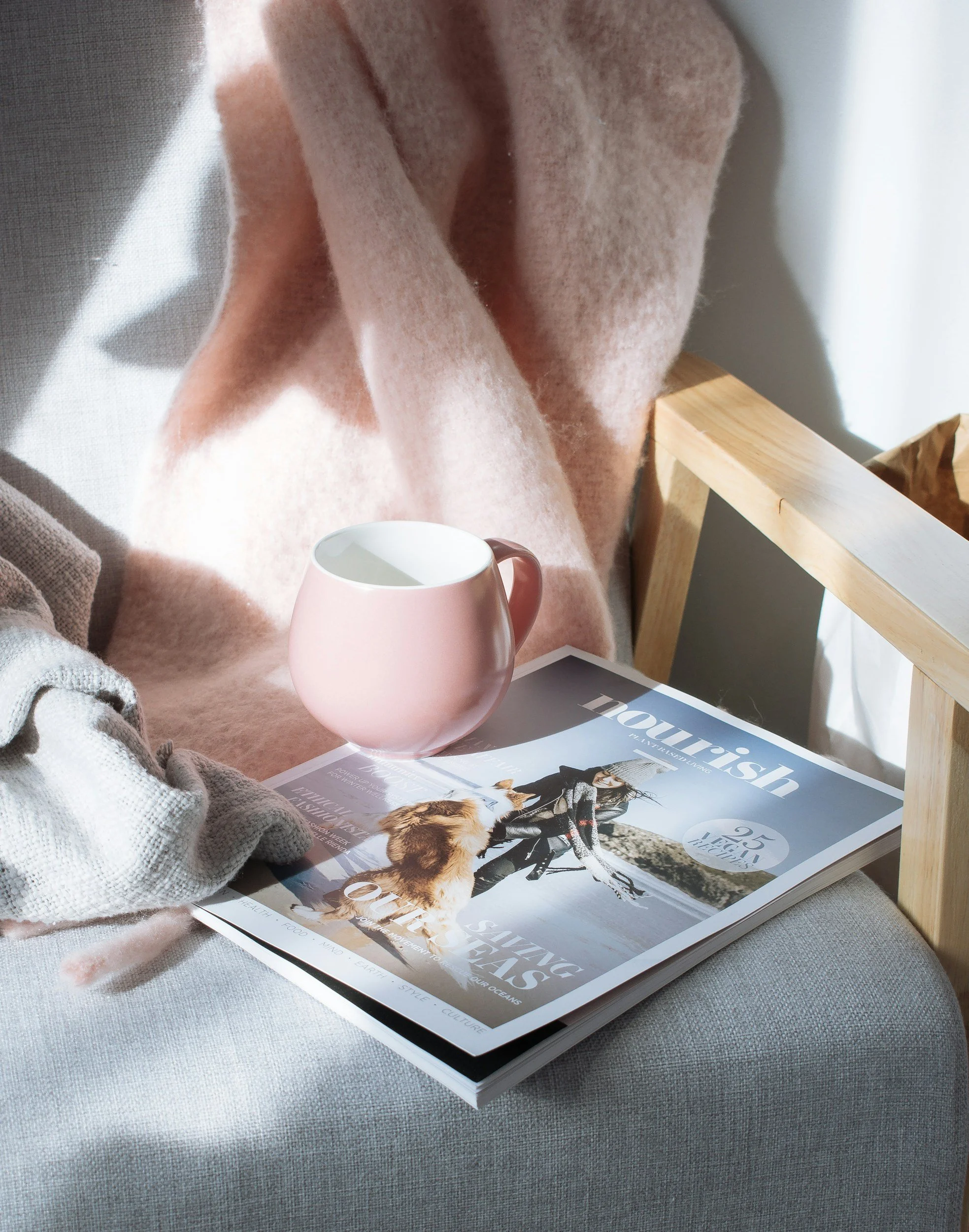 Ein rosa Kaffeebecher, eine Zeitschrift namens 'louvreish' mit einem Bild von jemandem, der auf einem Skiraum steht, und eine beige Decke auf einem grauen Stoffhintergrund. Auch ein Teil eines Holzrahmens ist sichtbar.