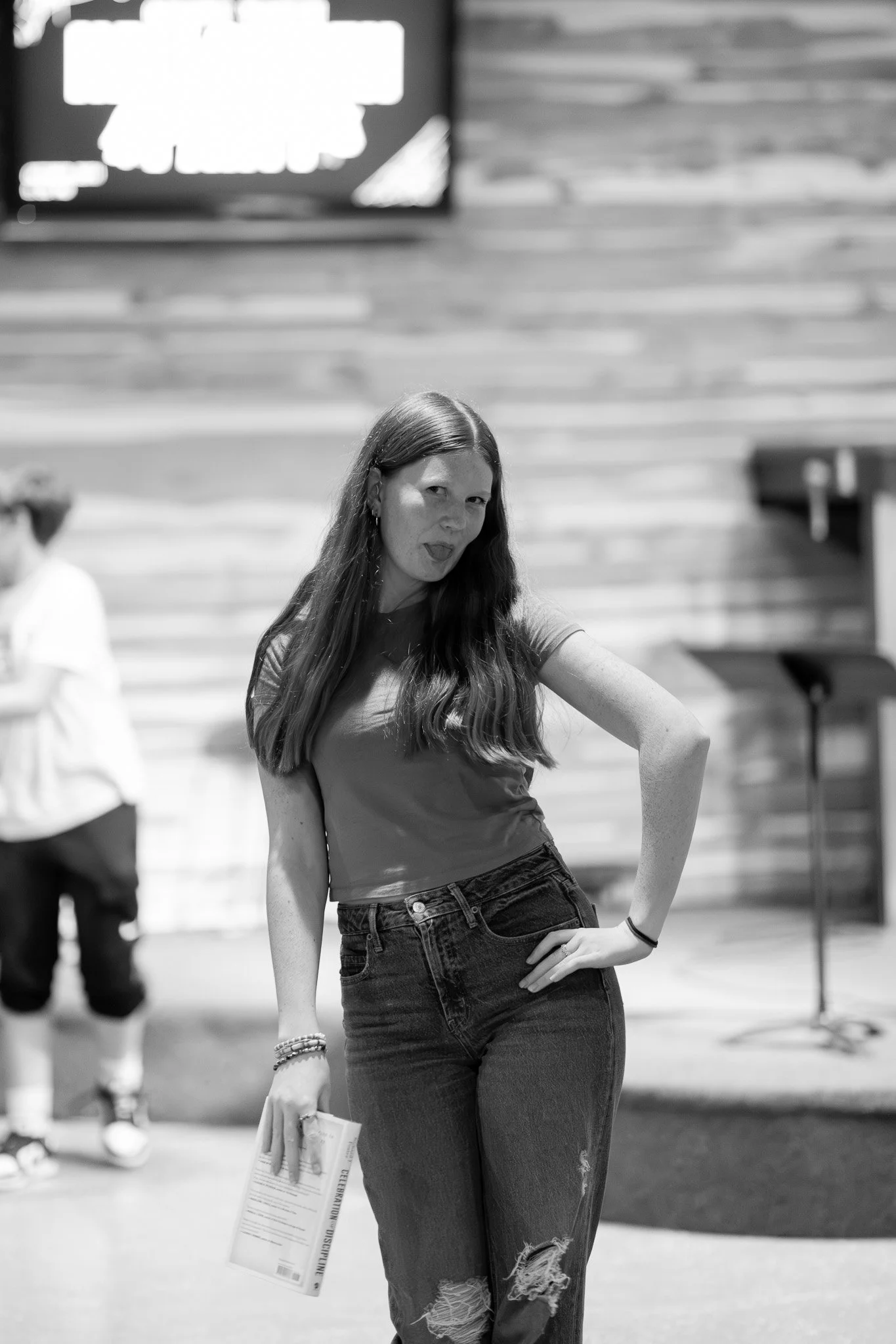 A young woman with long hair, wearing a t-shirt and ripped jeans, standing indoors and making a playful face.