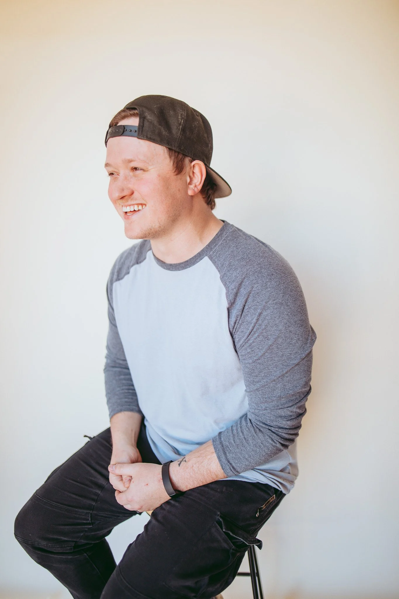A young man with a baseball cap backward, smiling and sitting on a stool against a plain white wall, dressed in a gray and white baseball-style shirt and black pants.