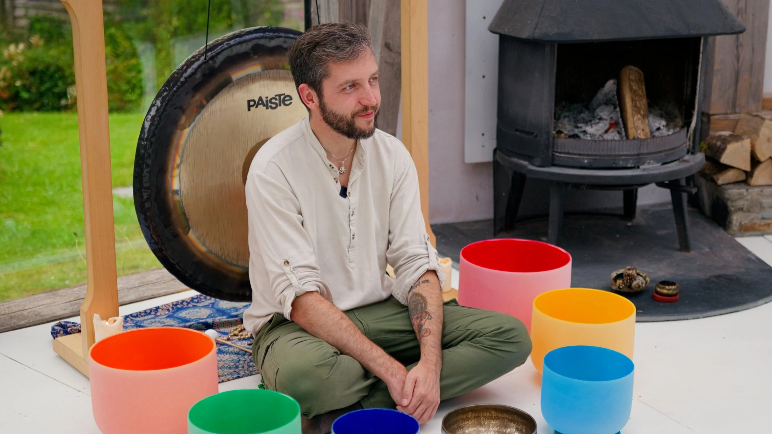 A man with a beard, dressed in white, sitting cross-legged on the floor playing colorful singing bowls with a wooden mallet in a softly lit room near a window.