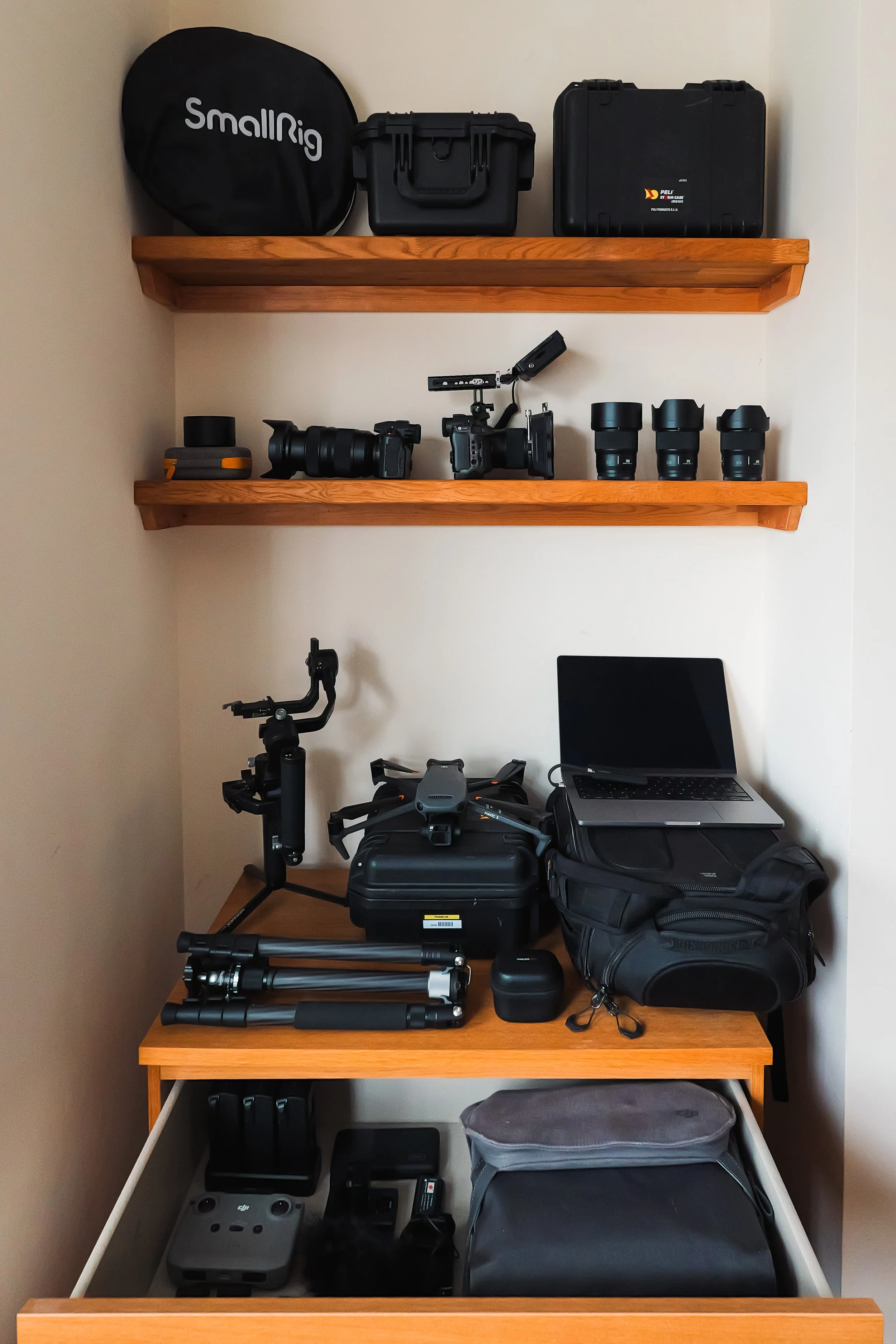 Organization of camera equipment and accessories on a wooden desk and shelves, including cameras, lenses, drone, tripod, cases, and a laptop.