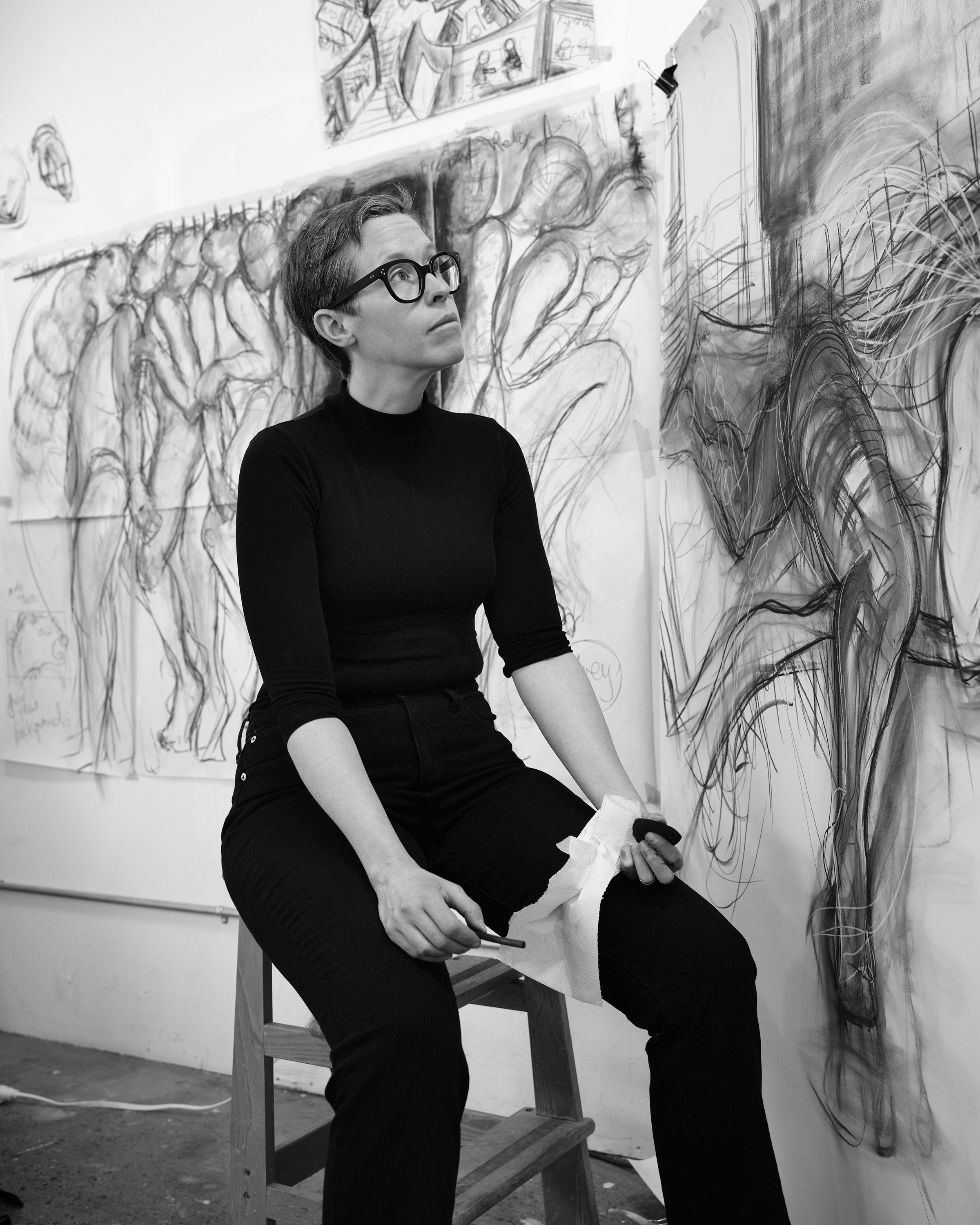 A woman with short hair and glasses sitting on a stool in front of a wall covered with sketches and drawings, holding a paintbrush and a tissue.