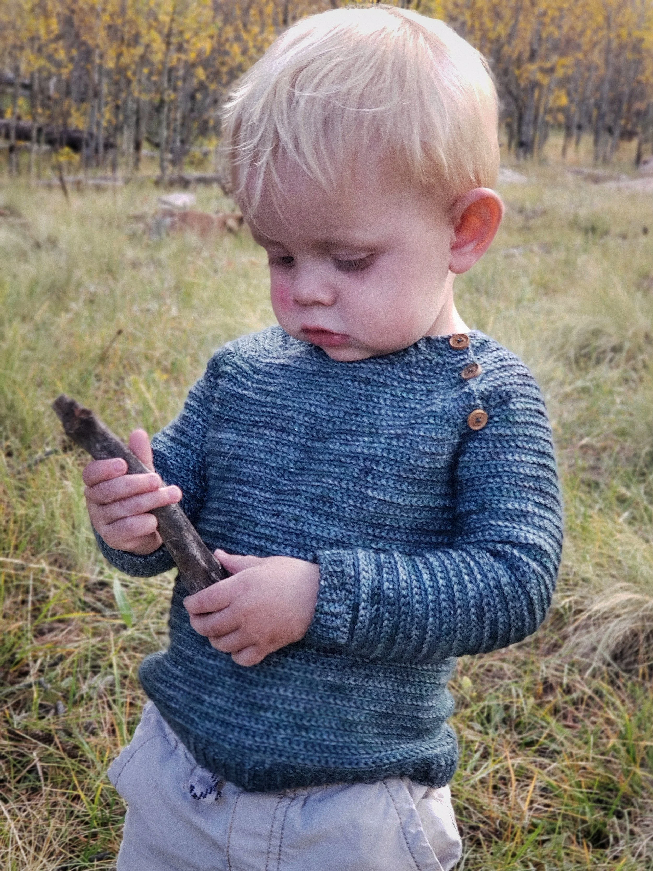 A green, handmade crochet raglan sweater on a young child, pictured from the front. The child examines a stick. The background is an aspen grove during late fall