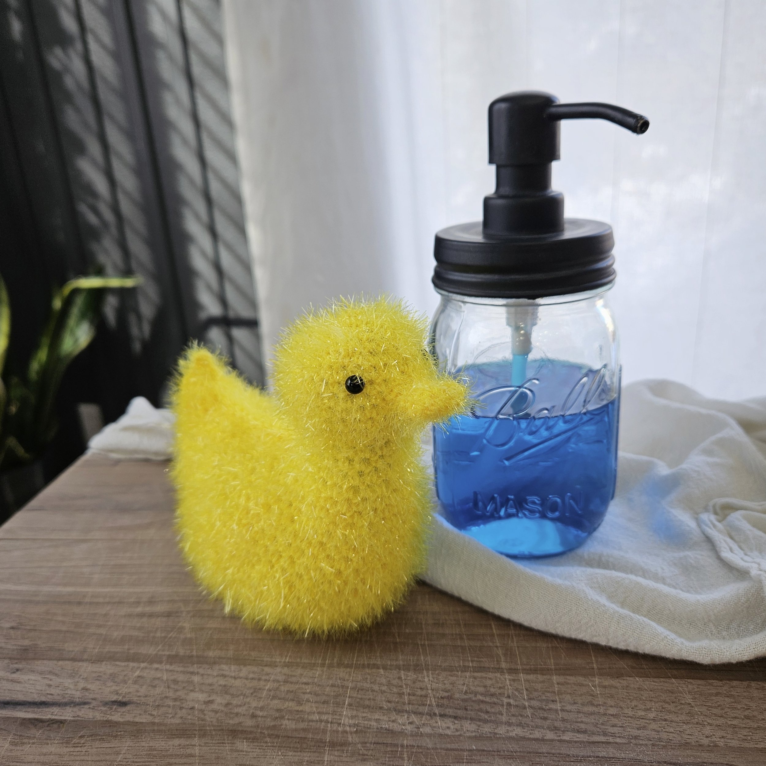 A fun and whimsical crochet duck-shaped scrubby made with scrubby yarn, perfect for washing dishes or gifting, placed next to a kitchen sink.