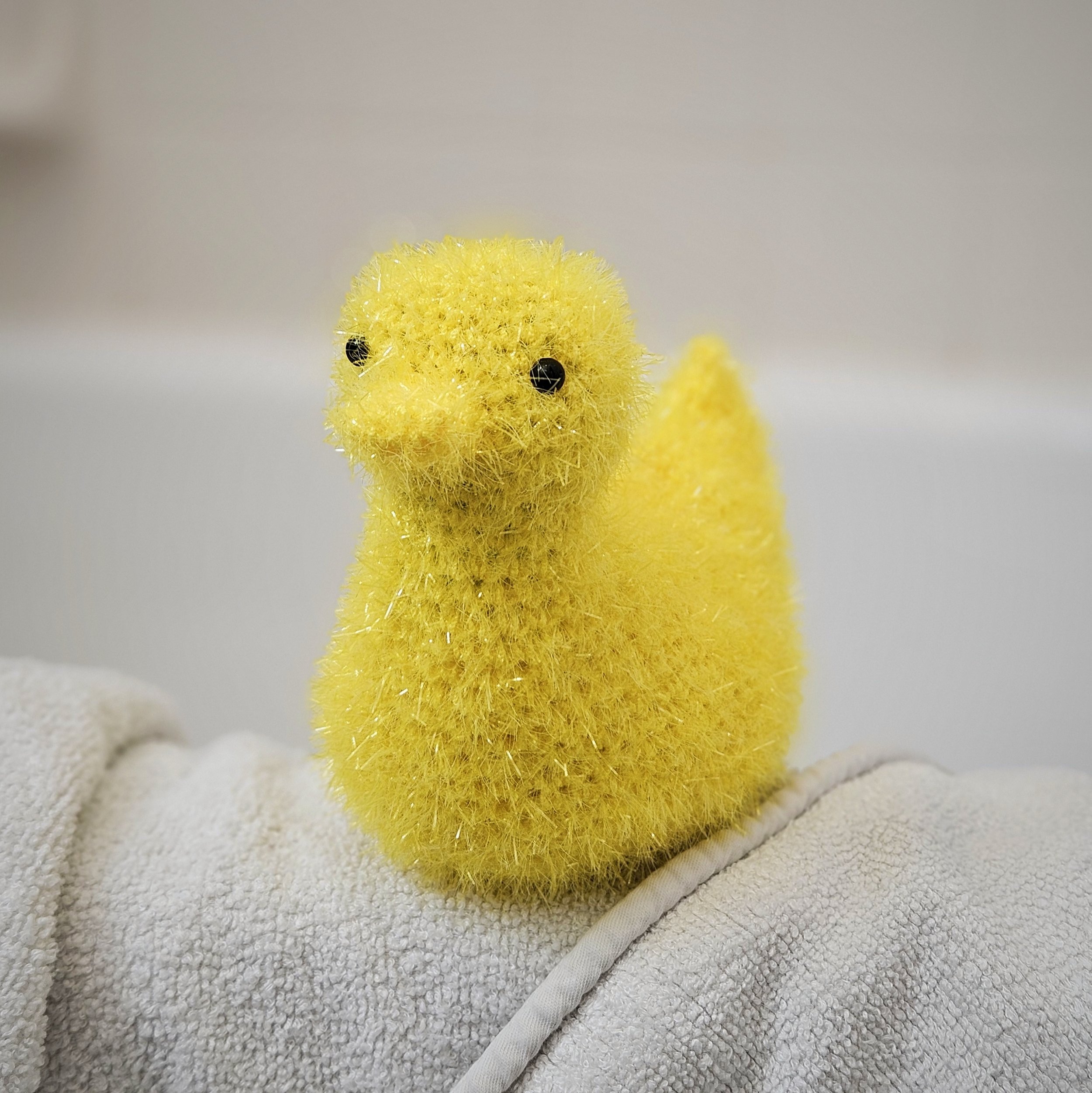 A cheerful crochet duck scrubby made with bright yellow scrubby yarn, sitting on the edge of a kitchen sink, ready for use.