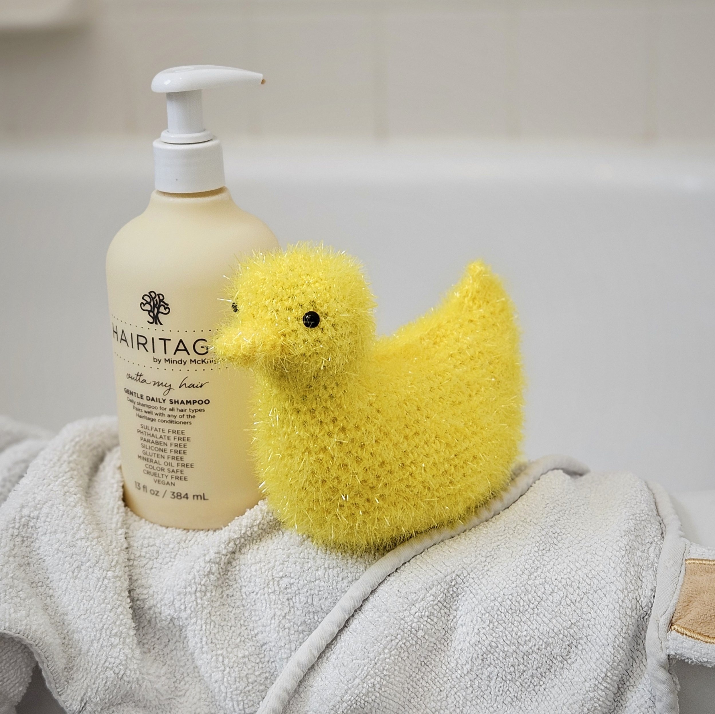 A crochet duck scrubby with soft, textured yarn, shown alongside a bottle of shampoo and a washcloth, highlighting its use for bath and body care.