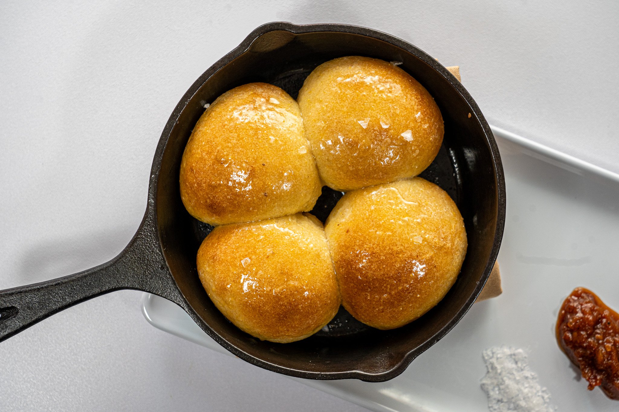 Four golden brown dinner rolls in a cast iron skillet with butter on top, accompanied by salt and a red sauce on a white plate.