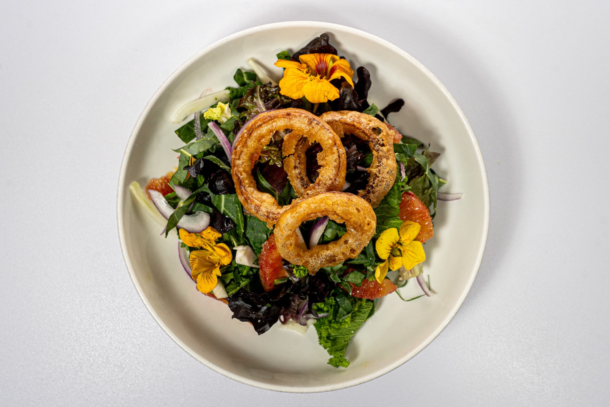 Salad with onion rings, mixed greens, edible flowers, red onion, and citrus slices in a white bowl.