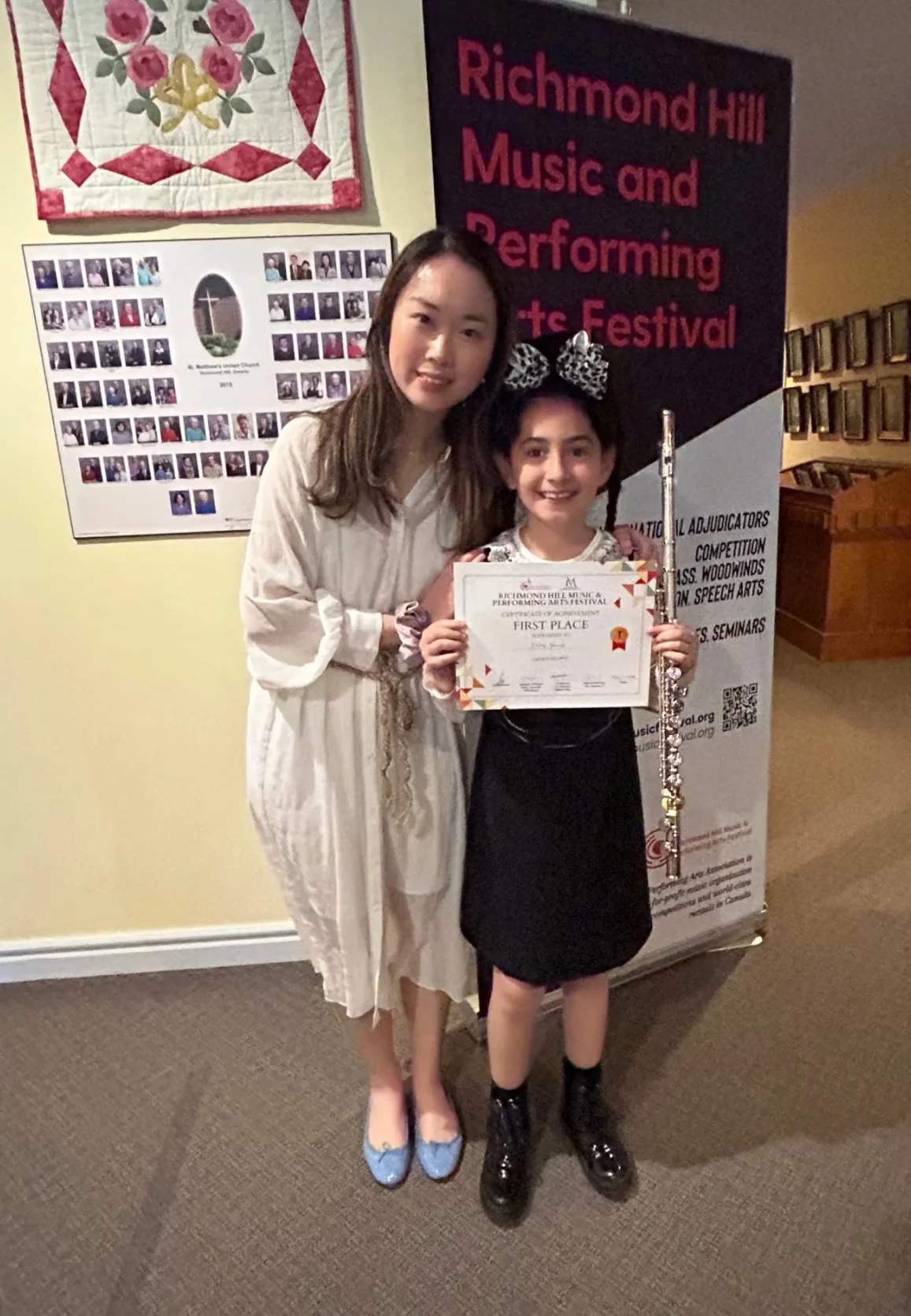 standing indoors at the Richmond Hill Music and Performing Arts Festival. One girl is holding a certificate and a flute. They are smiling and posing for the photo.