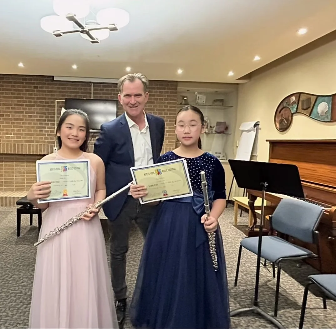 Two young girls in holding certificates and flutes, standing next to a man in a suit at a music competition
