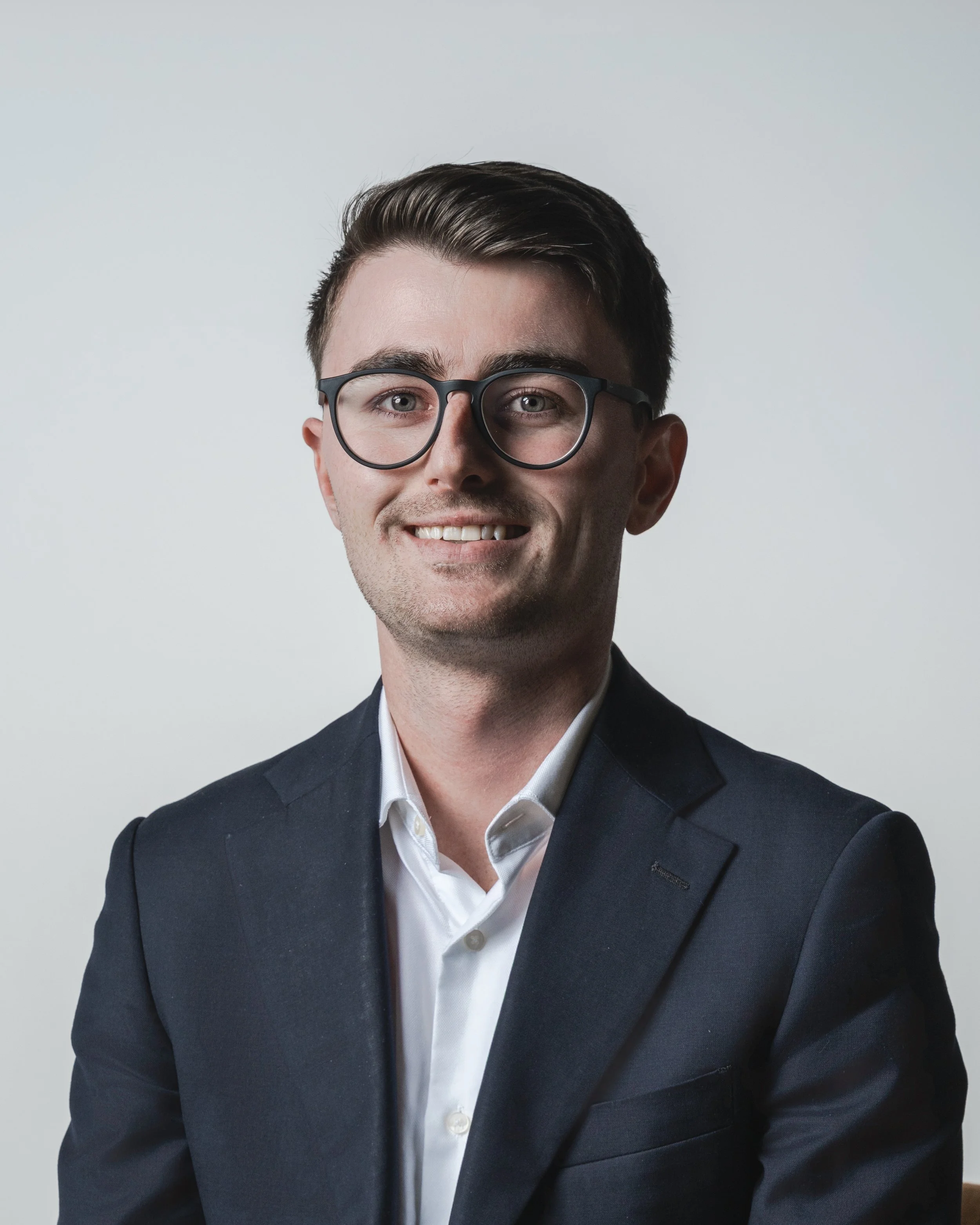 Corporate headshot of a young man in glasses and a navy suit on a grey background