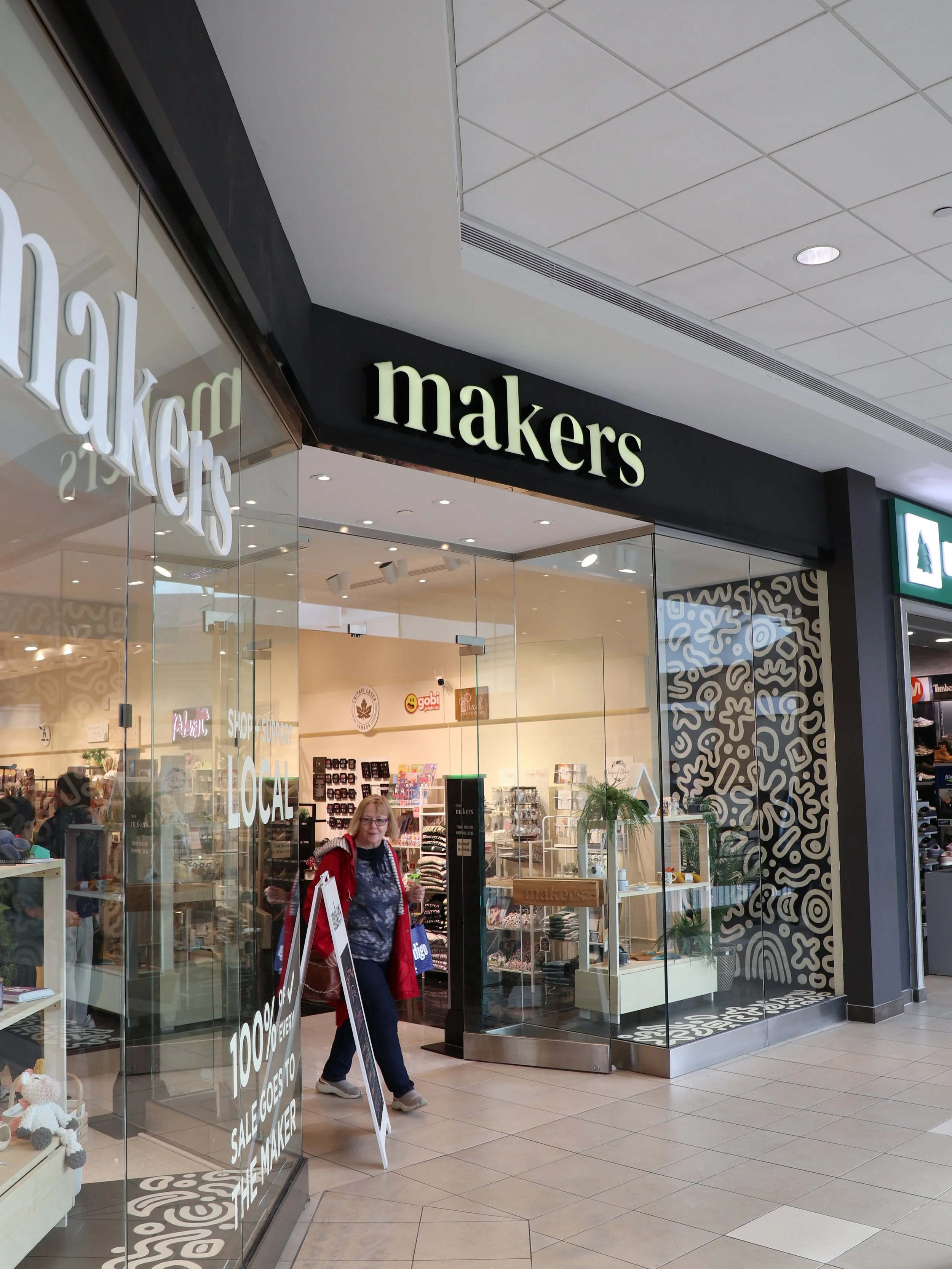 View of the storefront for the shop called 'makers' in a shopping mall, with a woman exiting the store carrying shopping bags.