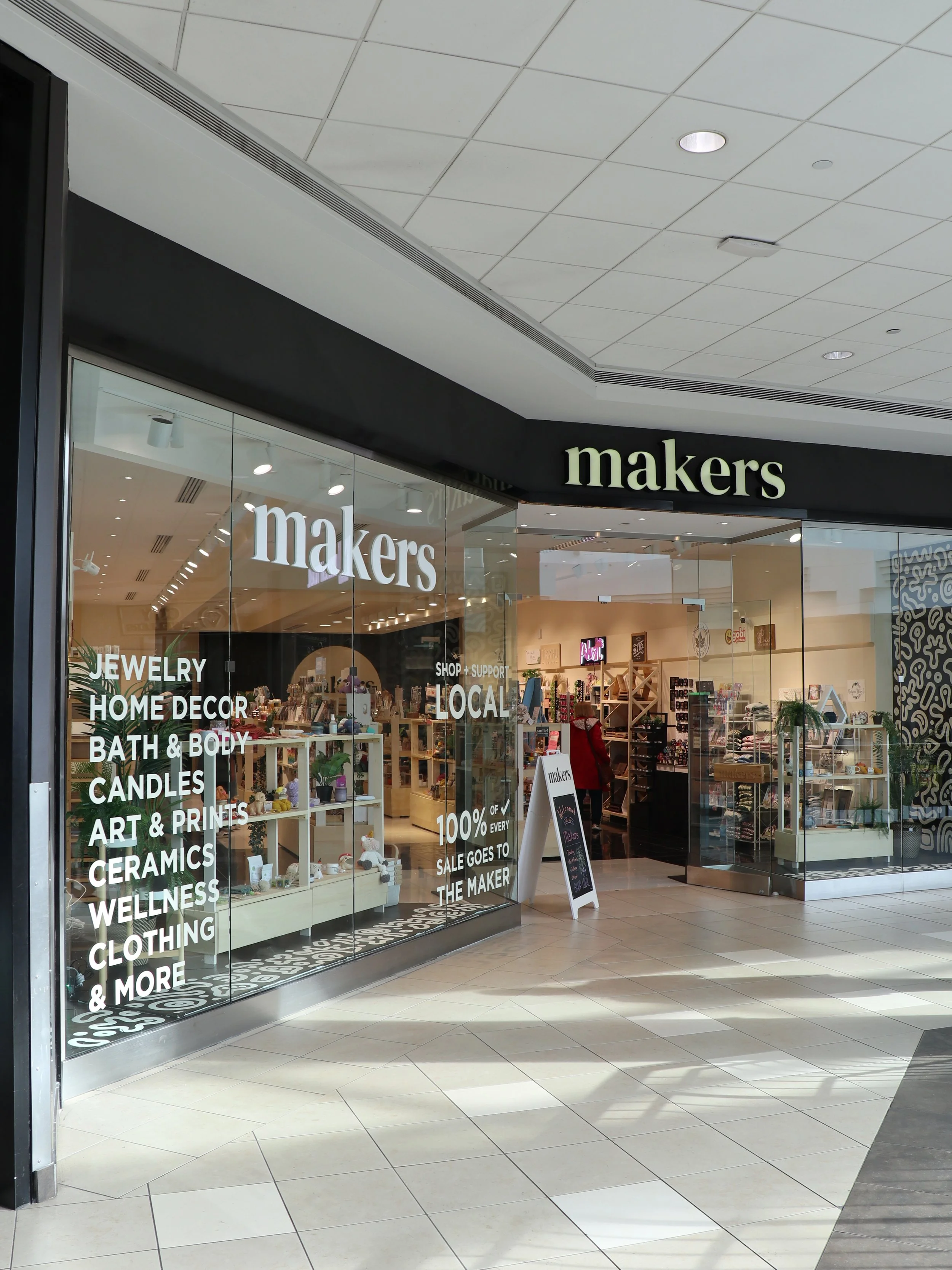 Entrance to a store named Makers selling jewelry, home decor, bath and body products, candles, art prints, ceramics, wellness, clothing, and more, with glass windows showcasing inside displays.