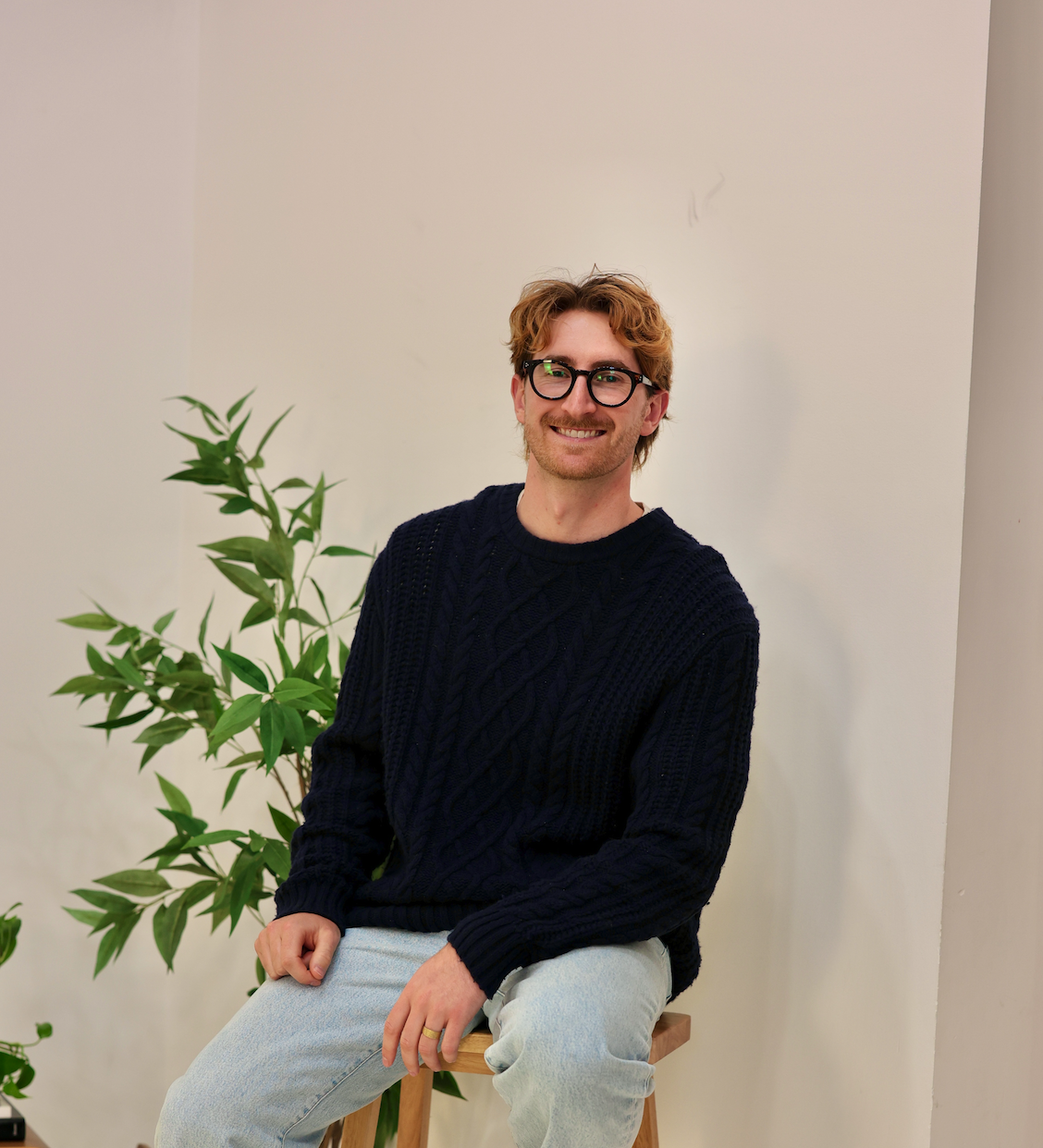 A smiling man with glasses, sitting on a wooden stool, wearing a navy blue sweater and light-colored pants, with a green plant behind him and a plain, off-white wall in the background.