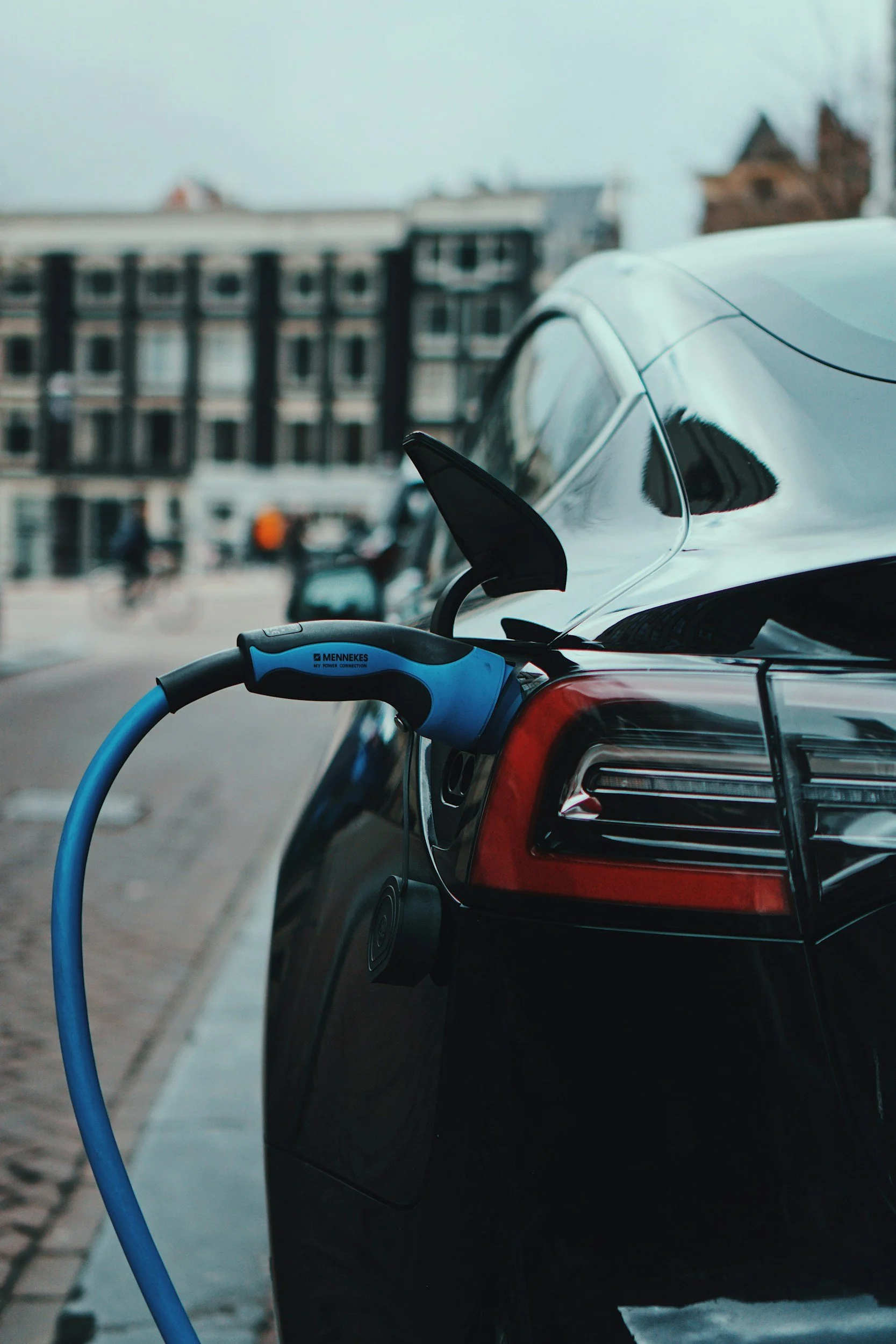 A close-up view of an electric vehicle charging station connected to a red car, parked on a street with residential buildings in the background.