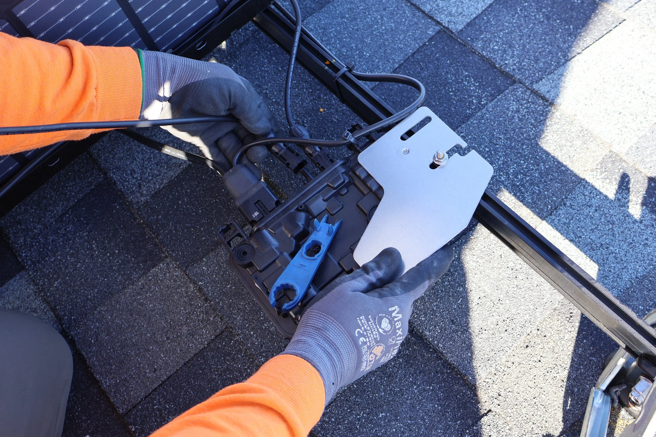 A person in gray gloves and orange sleeves working on a black solar panel or device on a rooftop. The person is using tools and cables, with the background showing gray asphalt shingles and part of a shadow.