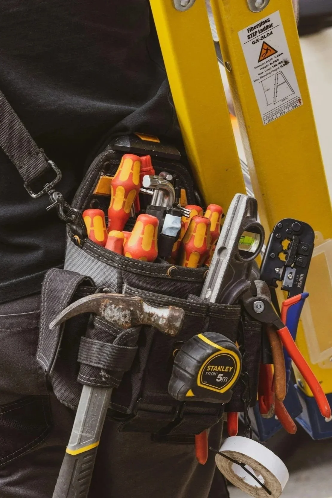 Close-up of a workman's tool belt containing various tools including screwdrivers, a hammer, a level, a pipe wrench, and a measuring tape. Part of a yellow ladder and a black bag are also visible.