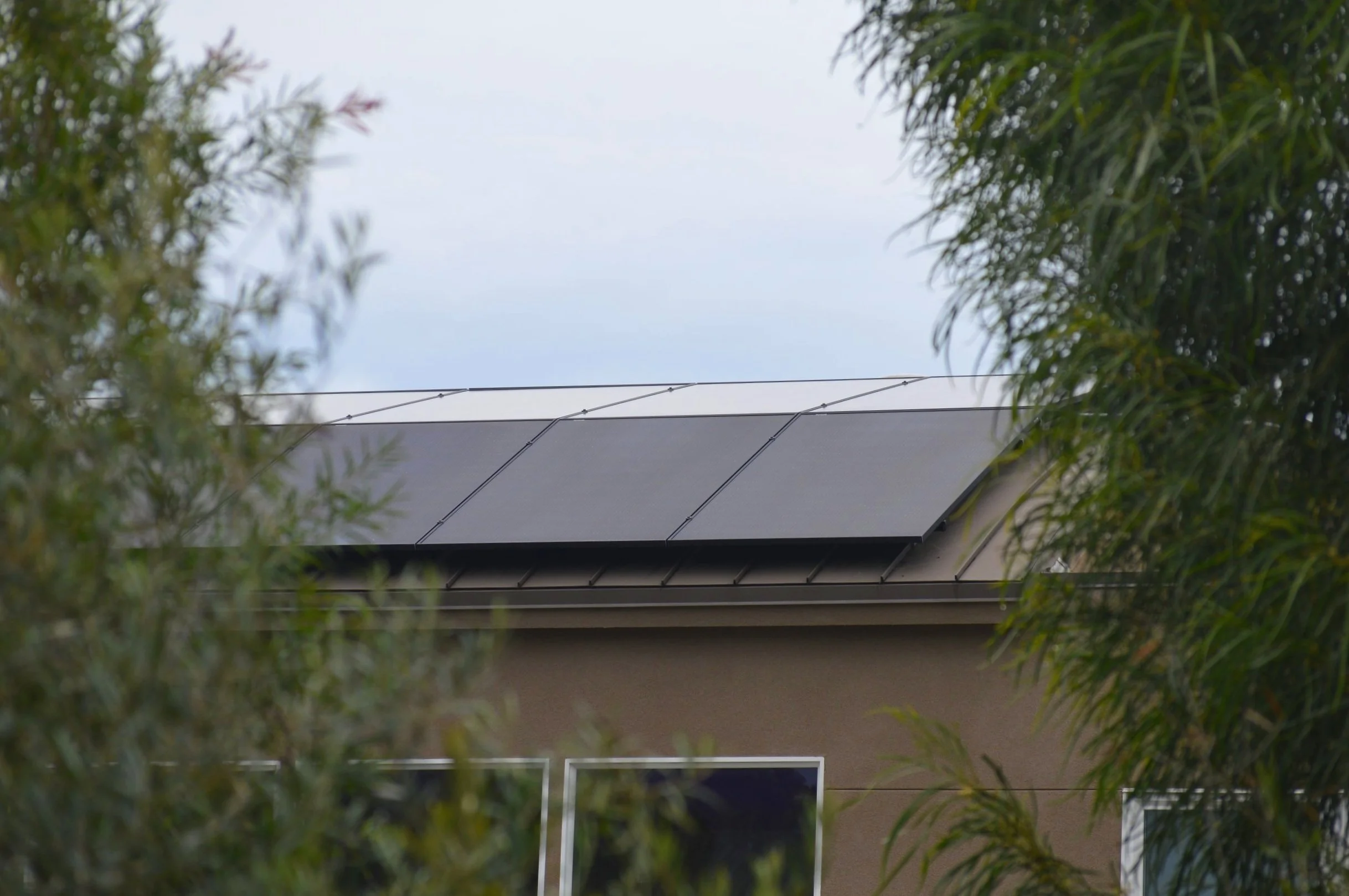 A house roof with solar panels, surrounded by trees and bushes, under a cloudy sky.