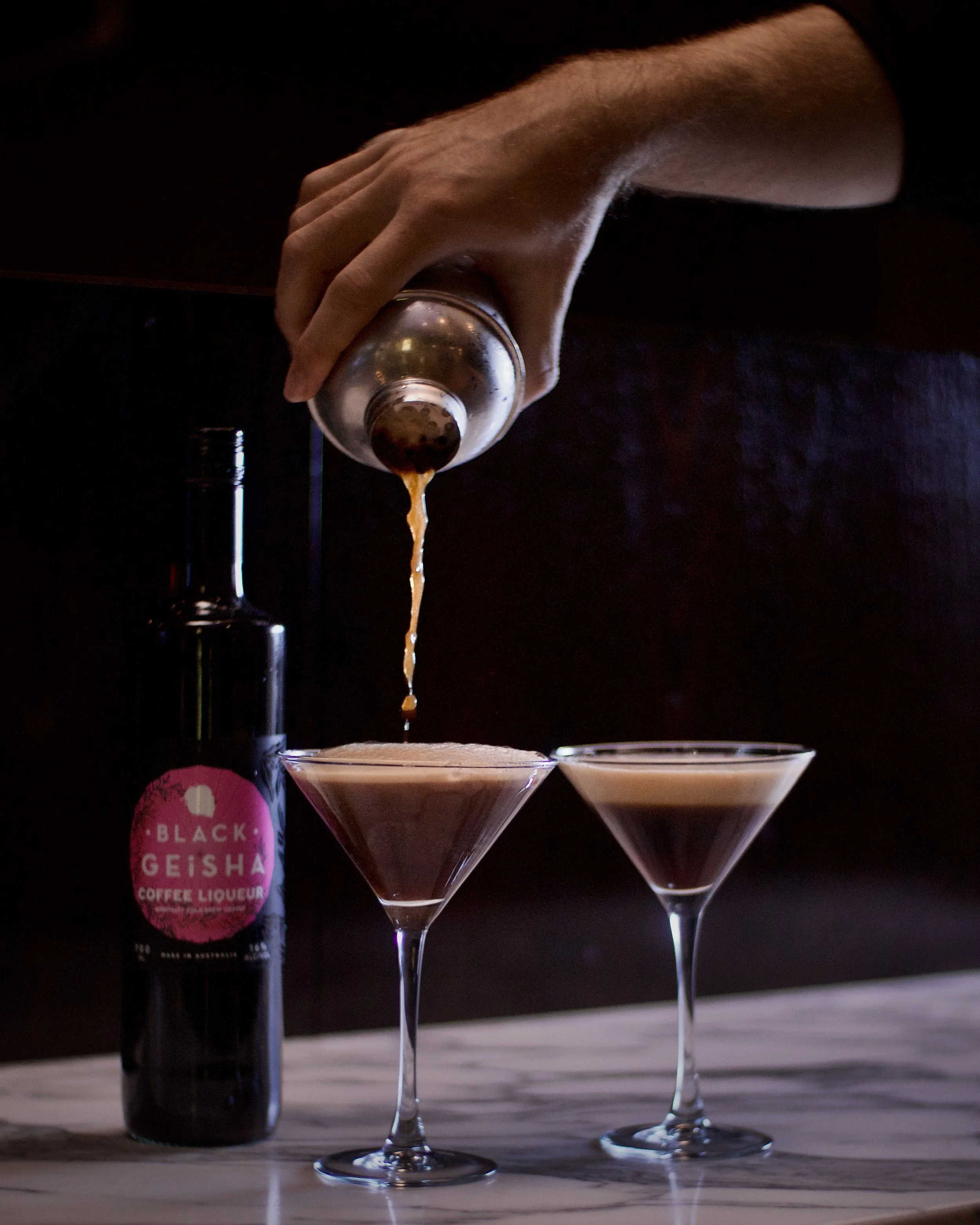 A hand pouring a coffee liqueur into a martini glass, with another similar glass next to it, on a marble surface, with a Black Geisha coffee liqueur bottle in the background.