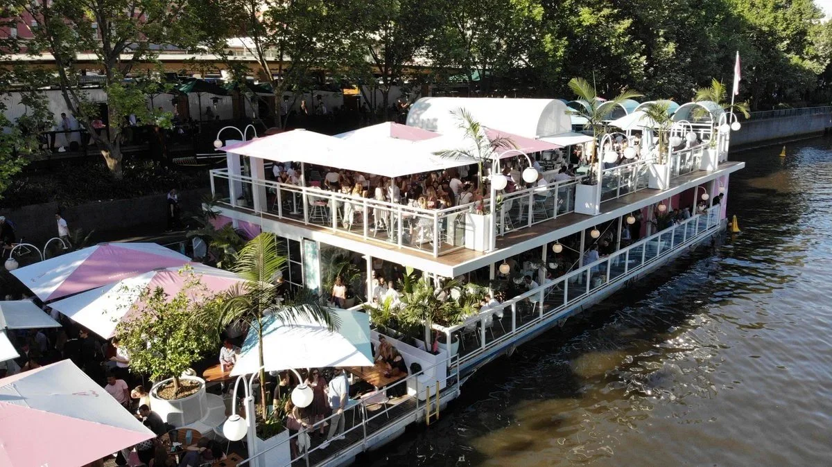 A large two-story boat docked on a river, converted into a restaurant with outdoor seating on both decks. The boat is decorated with pink and white umbrellas, tropical plants, and hanging lights. People are dining and socializing on both levels, with trees and a walkway visible on the riverbank.