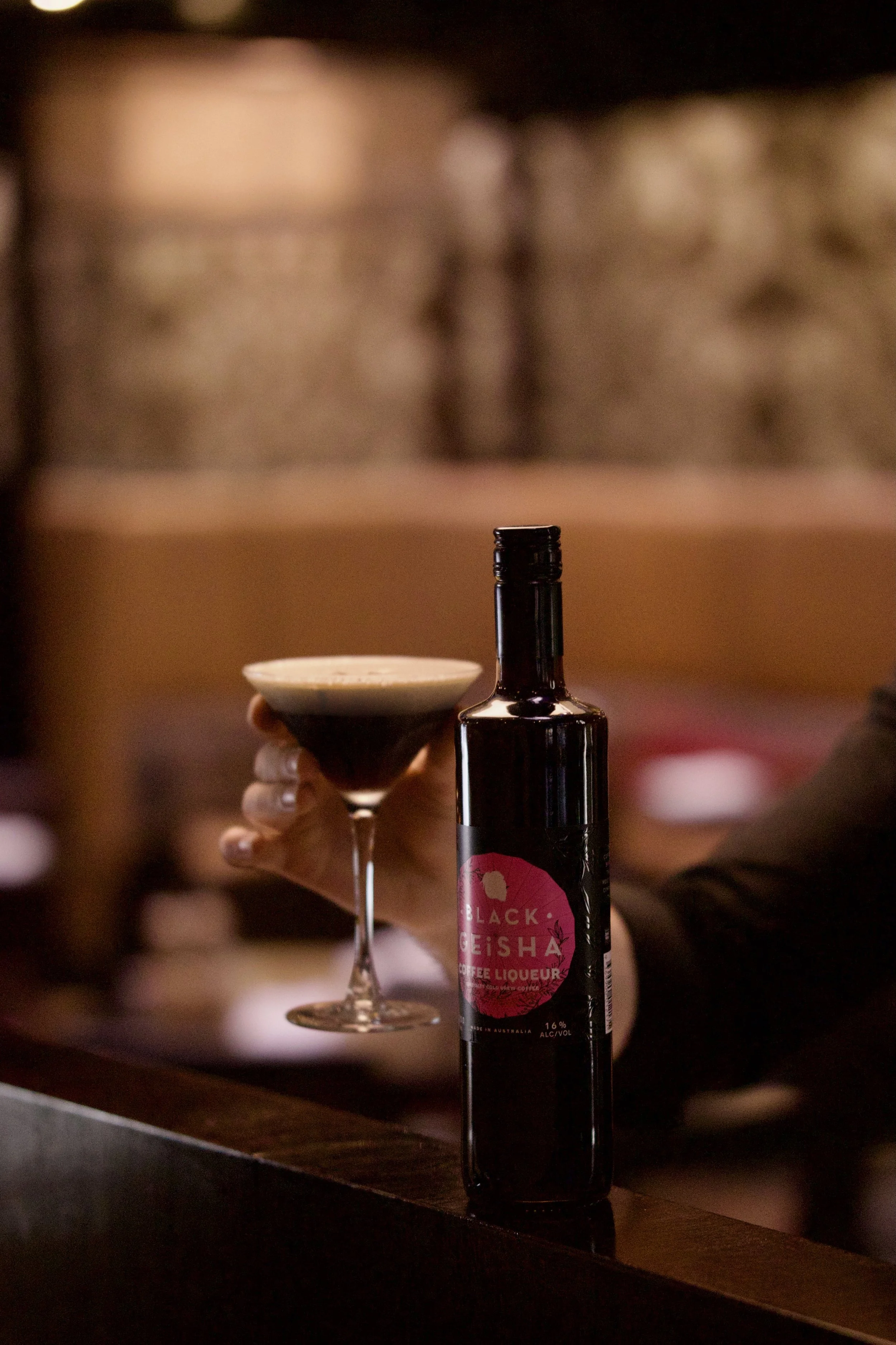 A hand holding a martini glass with a dark cocktail, next to a bottle of Black Bisha Coffee Liqueur on a bar counter.