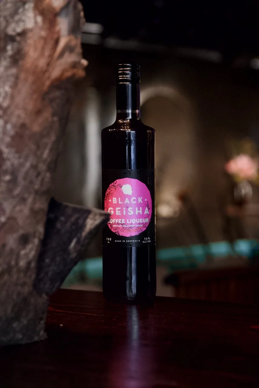 A dark bottle labeled 'Black Geisha Coffee Liqueur' with a pink and black design, placed on a wooden surface in a dimly lit setting.