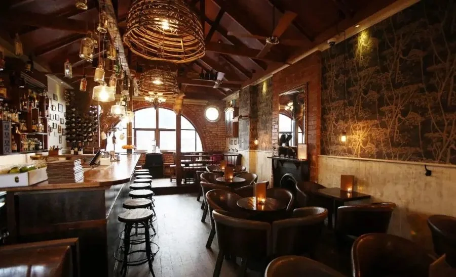 Interior of a cozy bar or restaurant with wooden furniture, brick walls, large arched windows, and warm lighting.