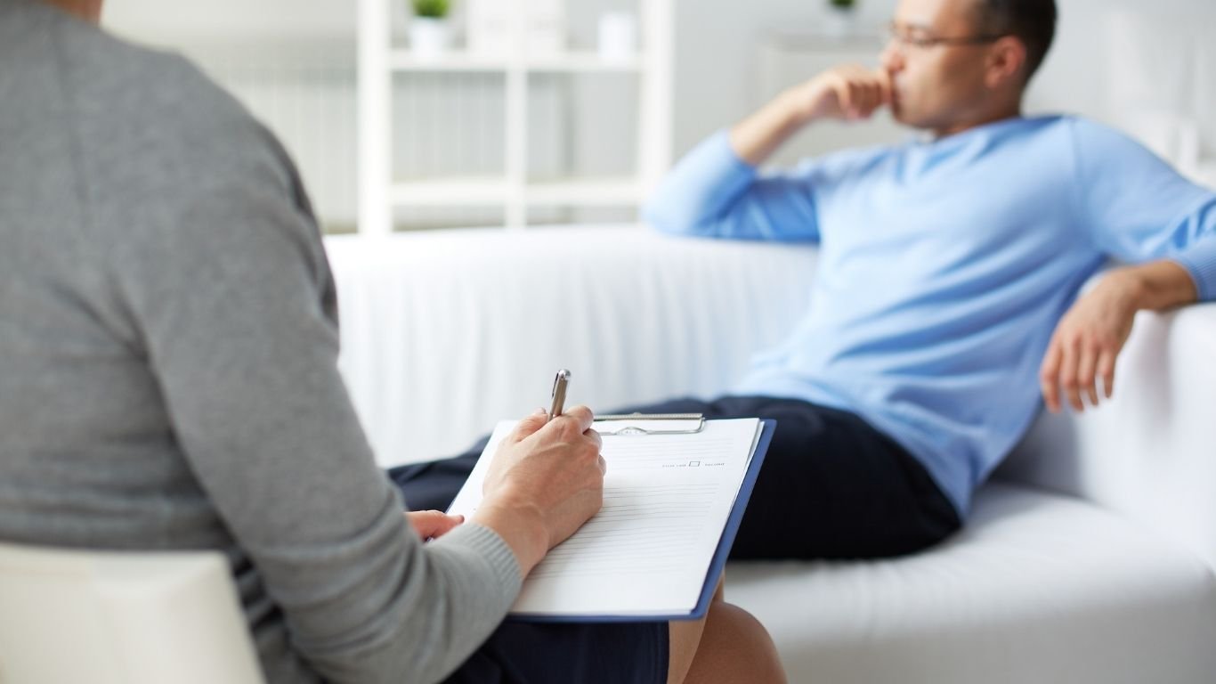 Therapist taking notes on a clipboard during a counseling session while a client sits on a couch in the background.