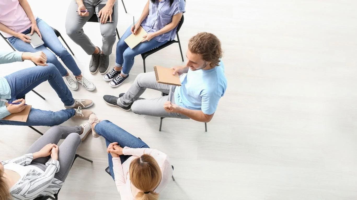 Group of adults seated in a circle holding notebooks, participating in a group counseling or discussion session.