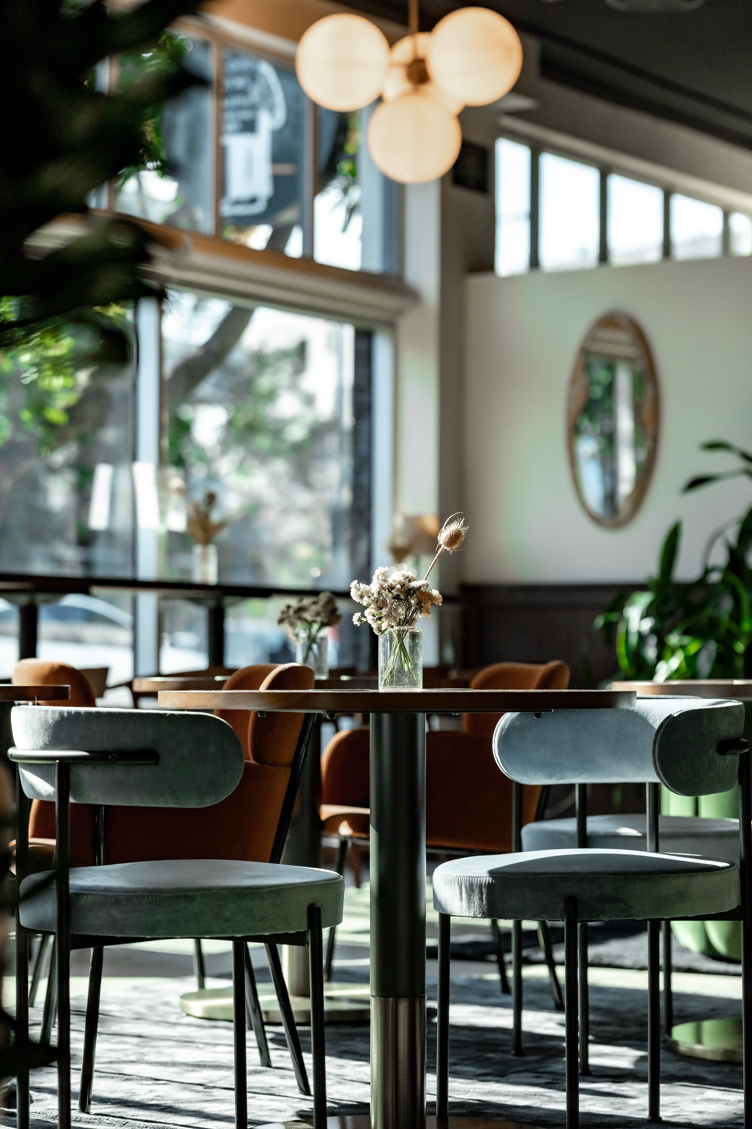 Empty restaurant with tables and chairs, decorated with flowers in vases, large windows, bright lighting, and modern interior design.