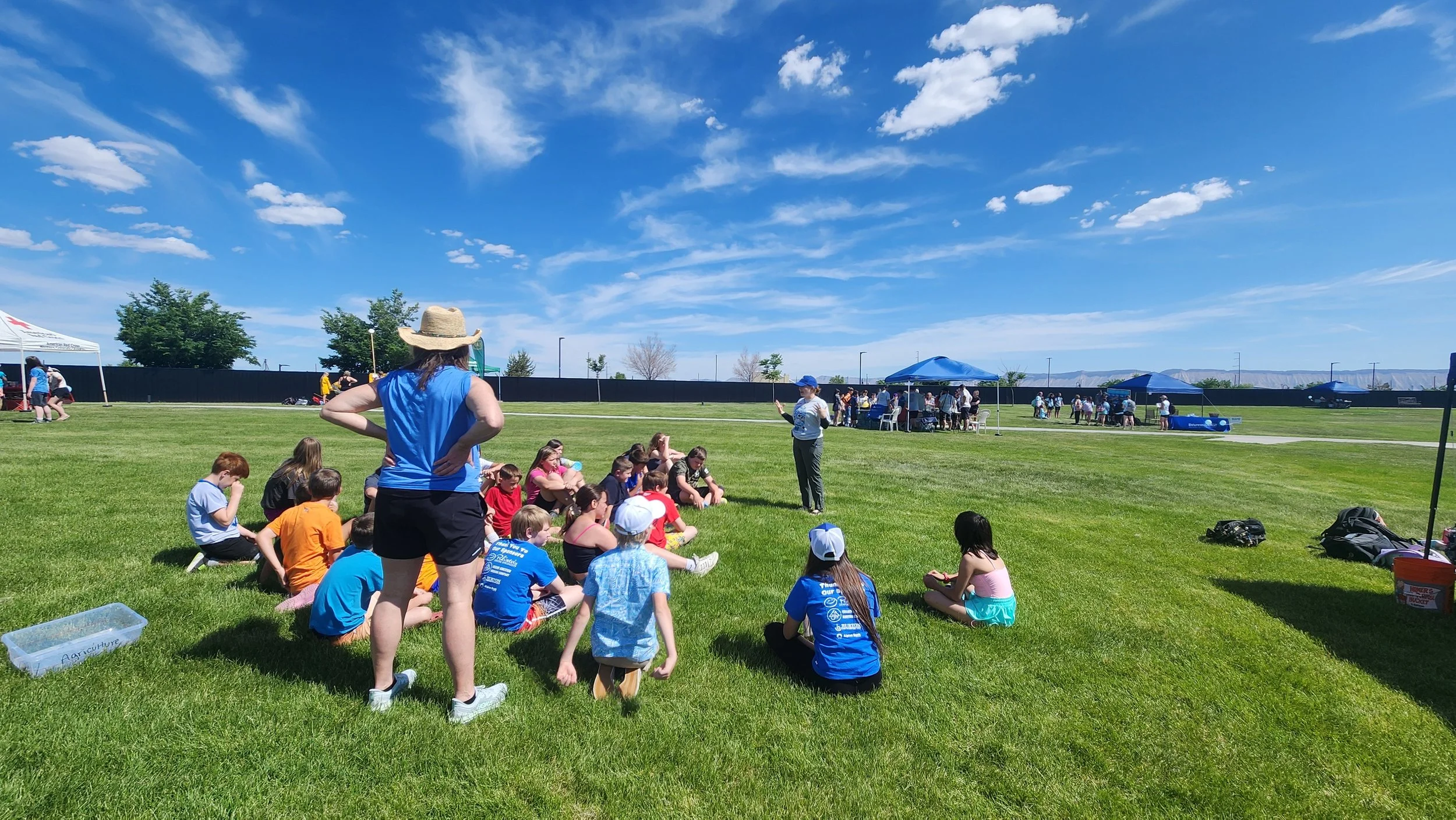 An adult is speaking to a group of students in a green grass field. Their teacher is standing behind them in a blue t-shirt and white brimmed hat