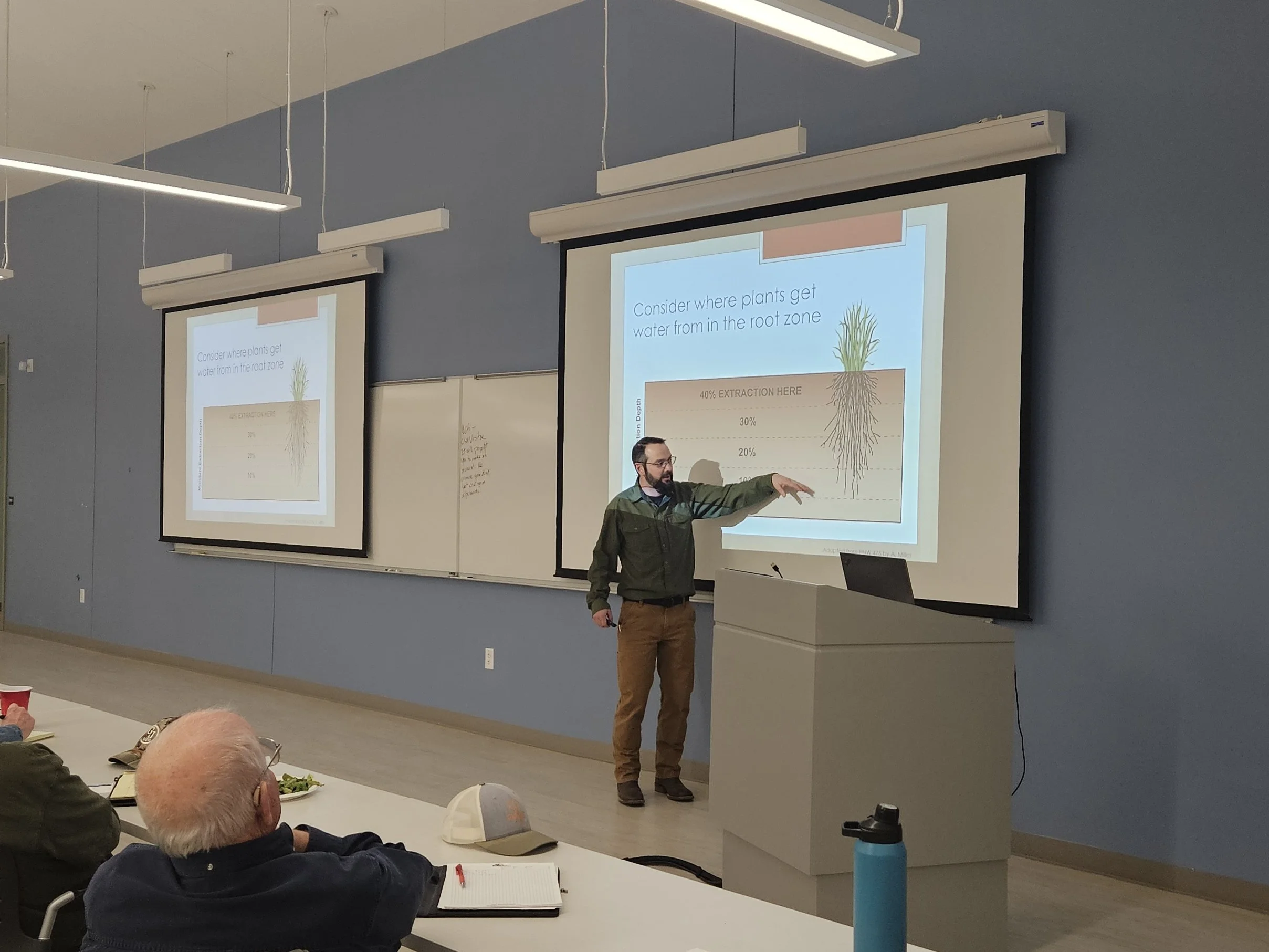 A man in a green shirt is presenting to a crowd and is pointing to a picture on a projection screen