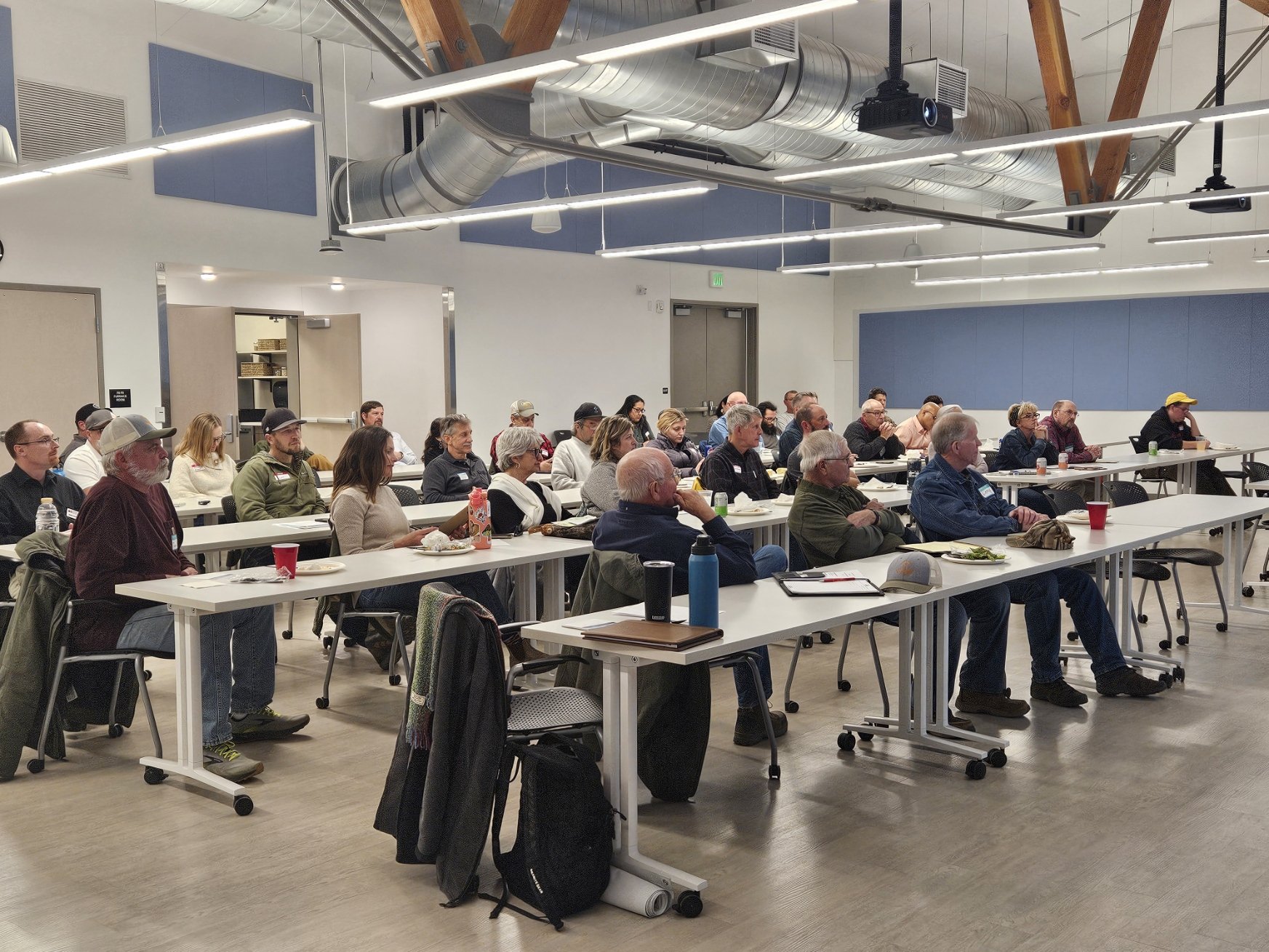 The crowd at the meeting is sitting in rows at white tables. They are looking at the front of the room