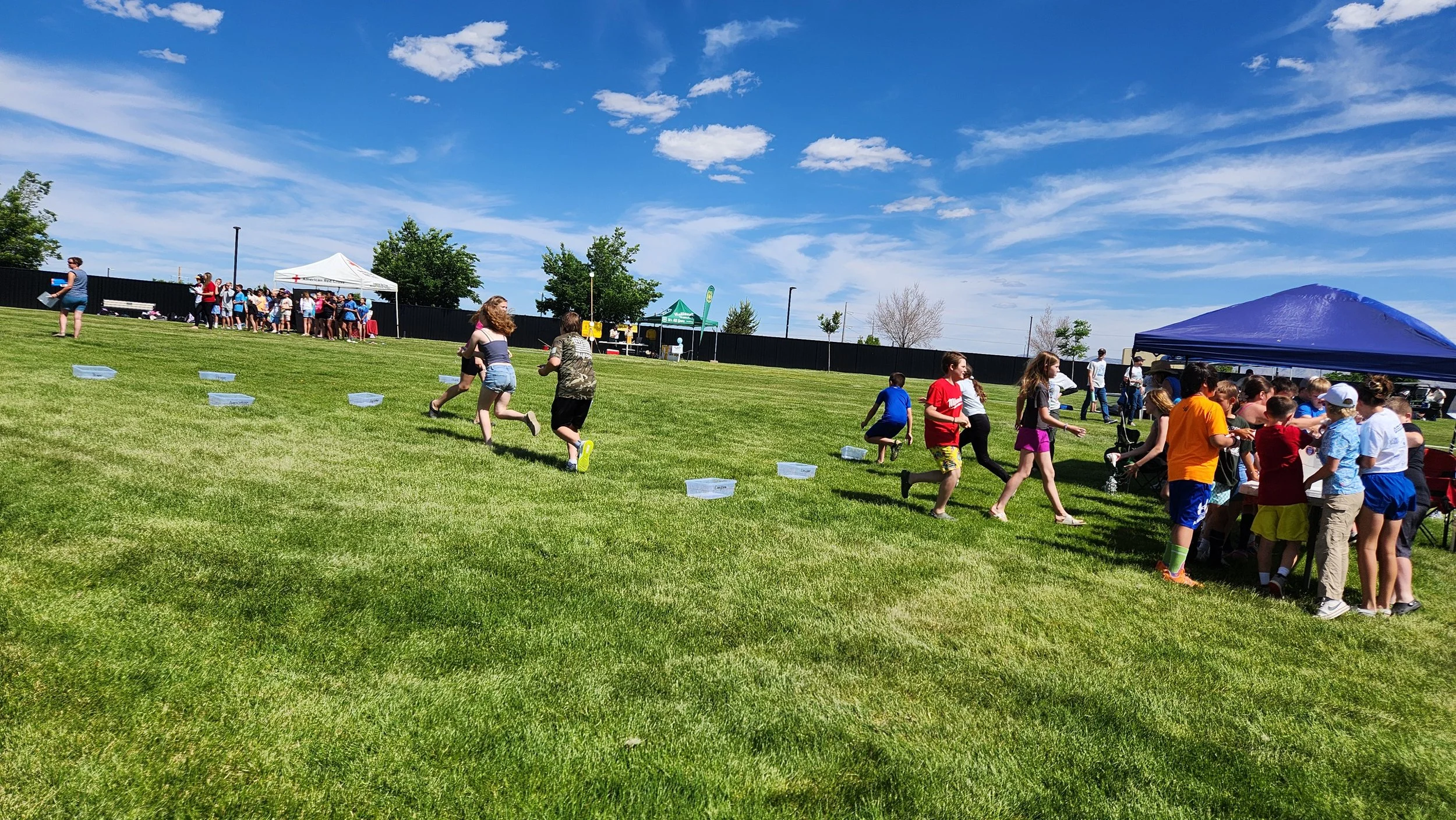 Students are running between clear bins in a green grassy field with a blue canopy in the background.