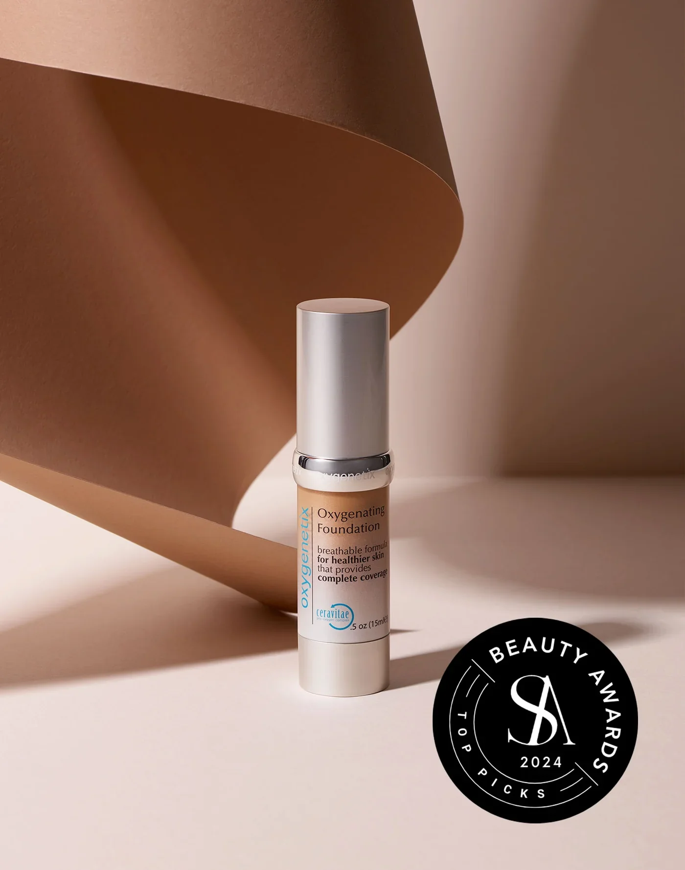 A bottle of Oxygenating Foundation with a silver cap, standing upright on a beige surface with a brown paper background, featuring a black circular beauty awards sticker for 2024.
