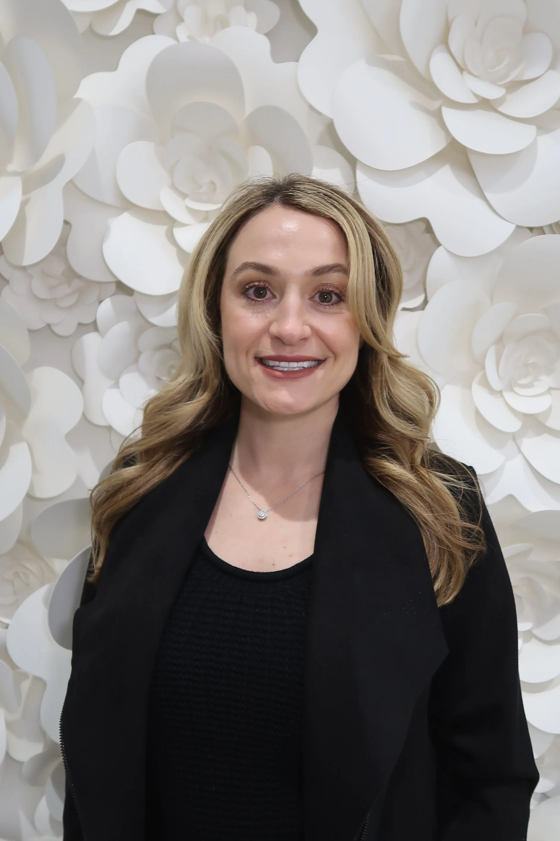 A woman with long, wavy blonde hair smiling, wearing a black top and a black jacket, standing in front of a decorative white floral background.