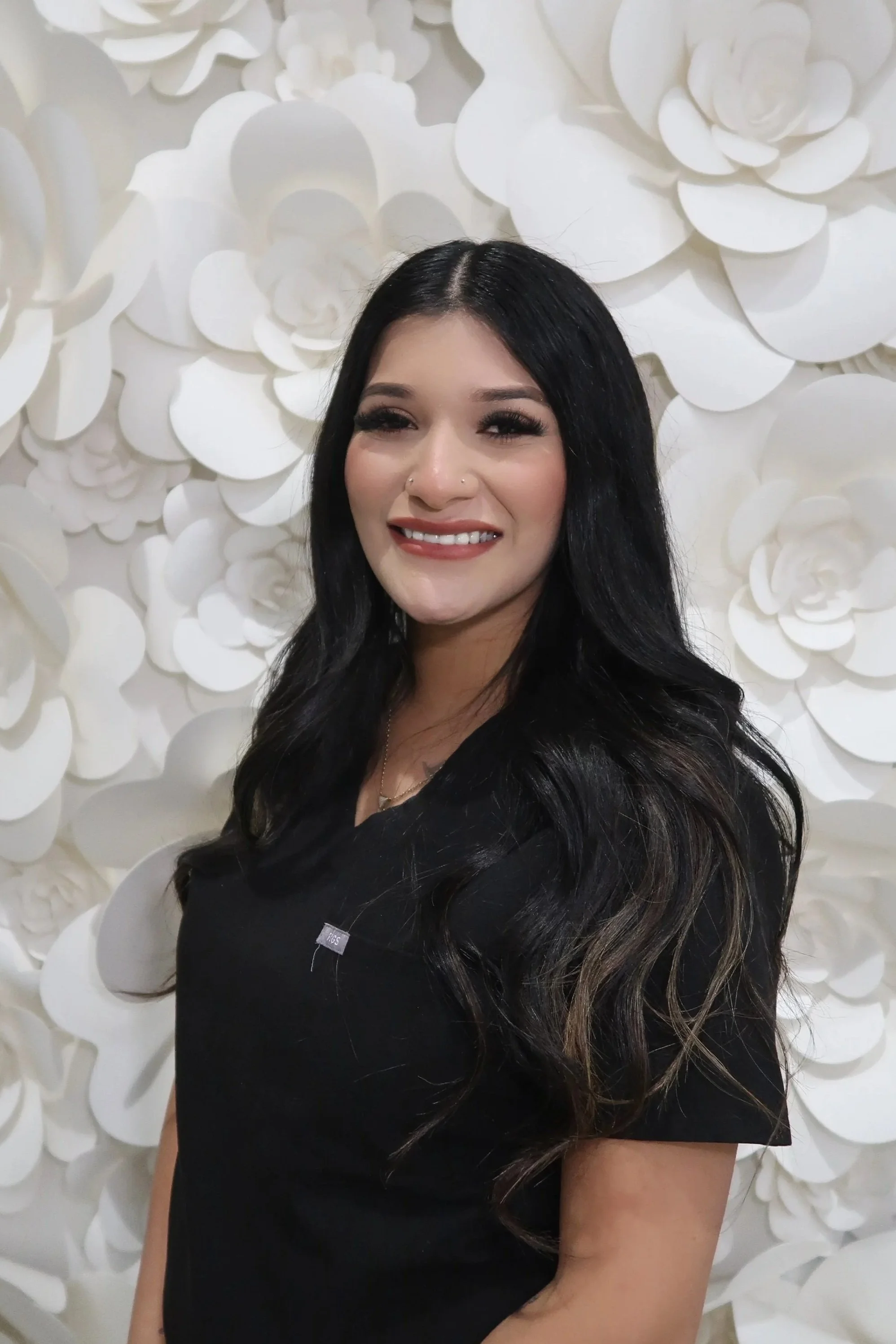 A woman with long, straight brown hair smiling in front of a white floral background, wearing a black blazer, a black turtleneck, and a cross necklace.