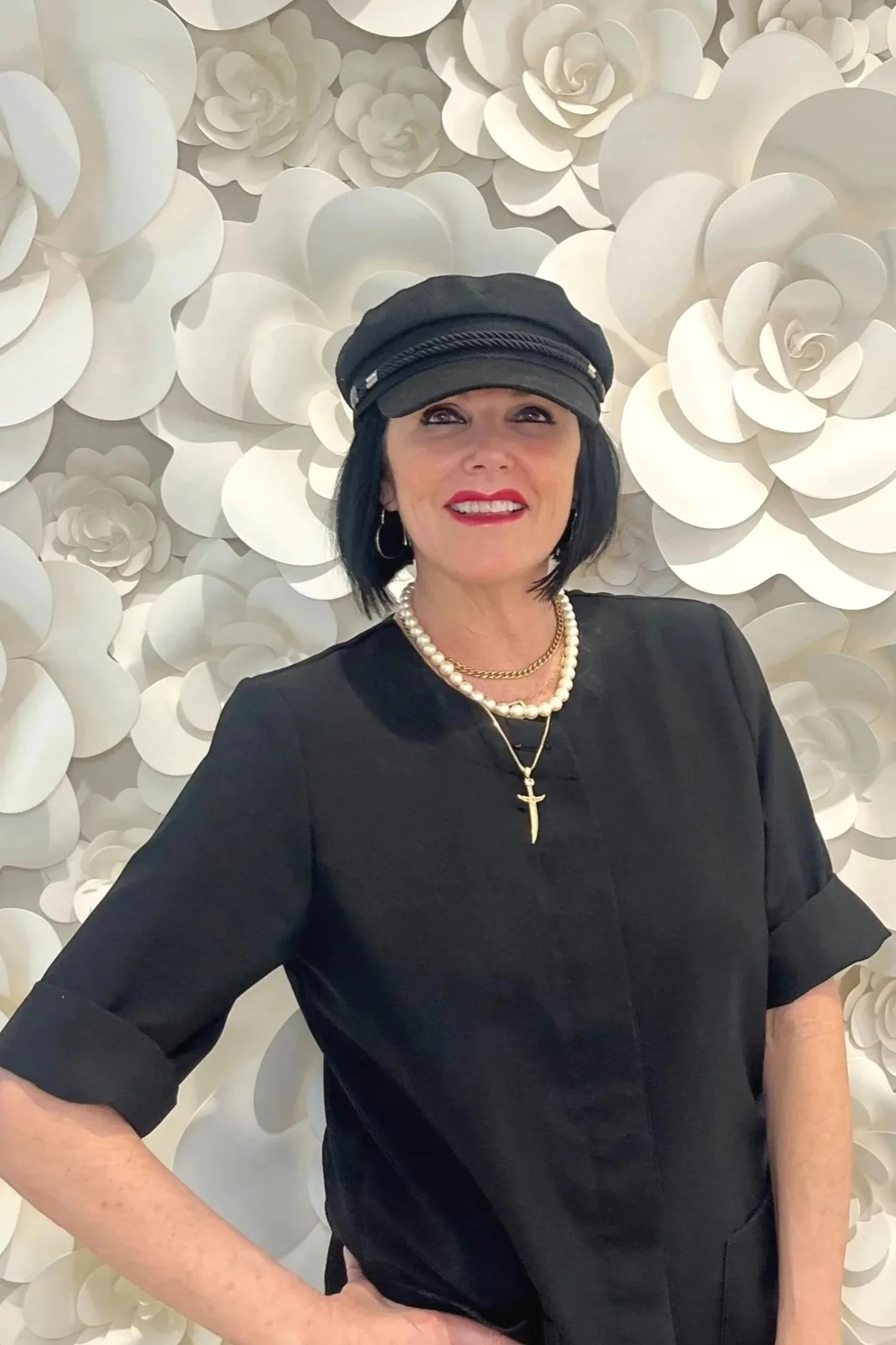 A woman with shoulder-length black hair wearing a black cap, black dress, pearl necklace, gold chain, and a gold cross pendant stands in front of a white floral wall.