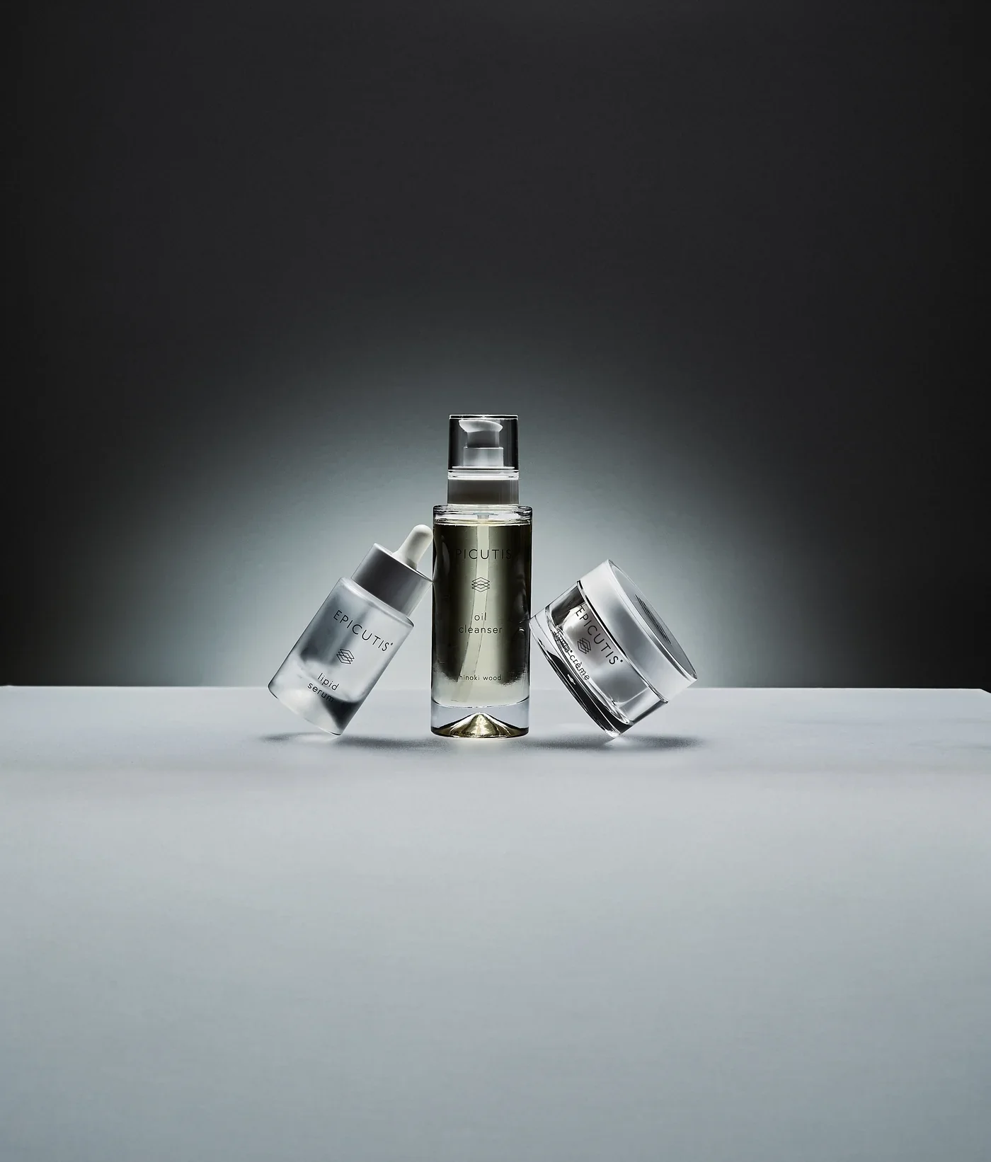A collection of three skincare products on a white surface against a gray gradient background. The products include a dropper bottle, a pump bottle, and a jar, all labeled with the brand Epicutis.