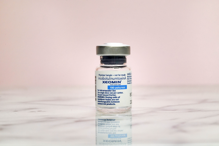 A small glass vial of Xeomin, a prescription medication, on a marble surface with a light pink background.