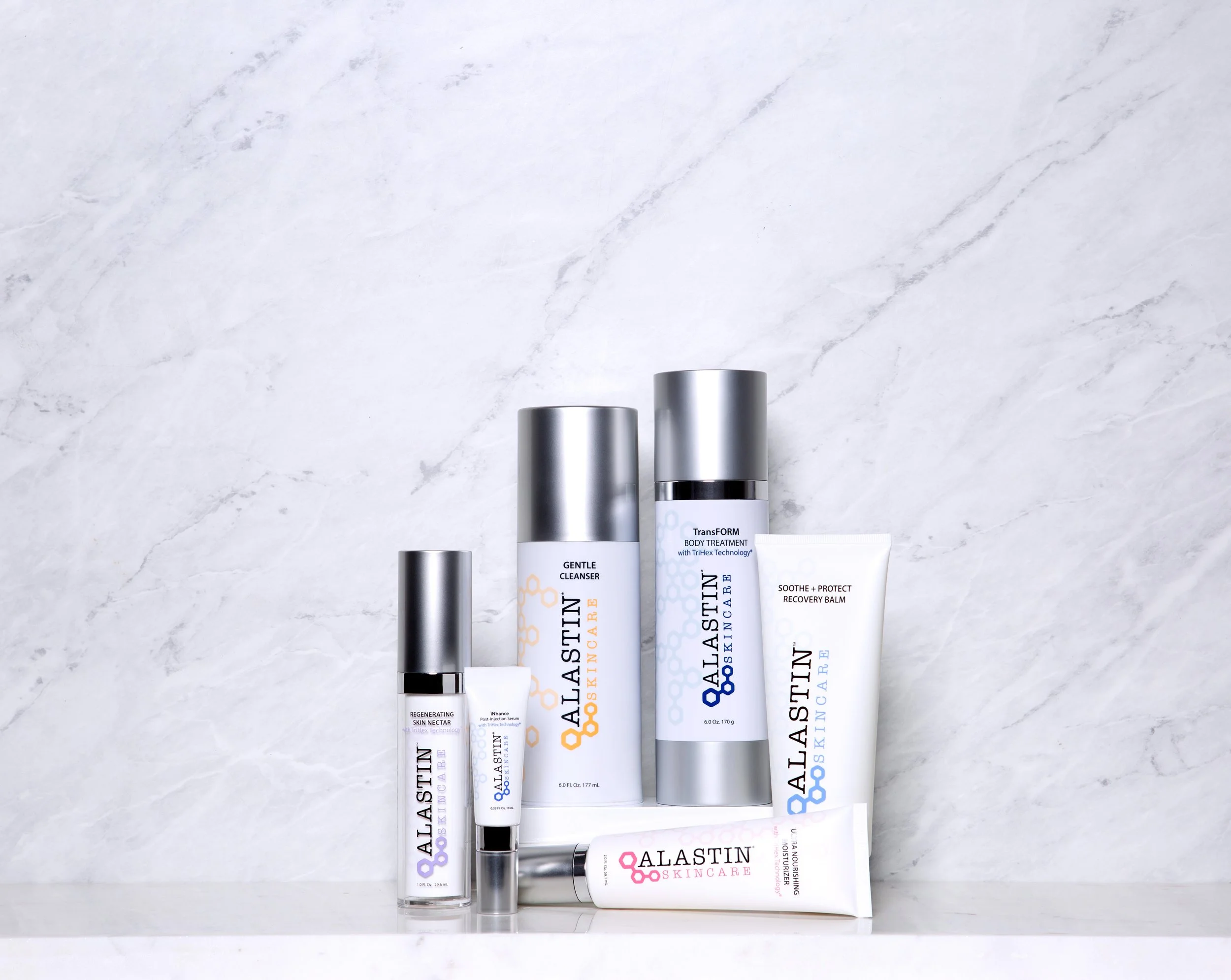 Collection of skincare products on a white marble surface, including bottles and tubes labeled Alastin Skincare, against a white marble background.