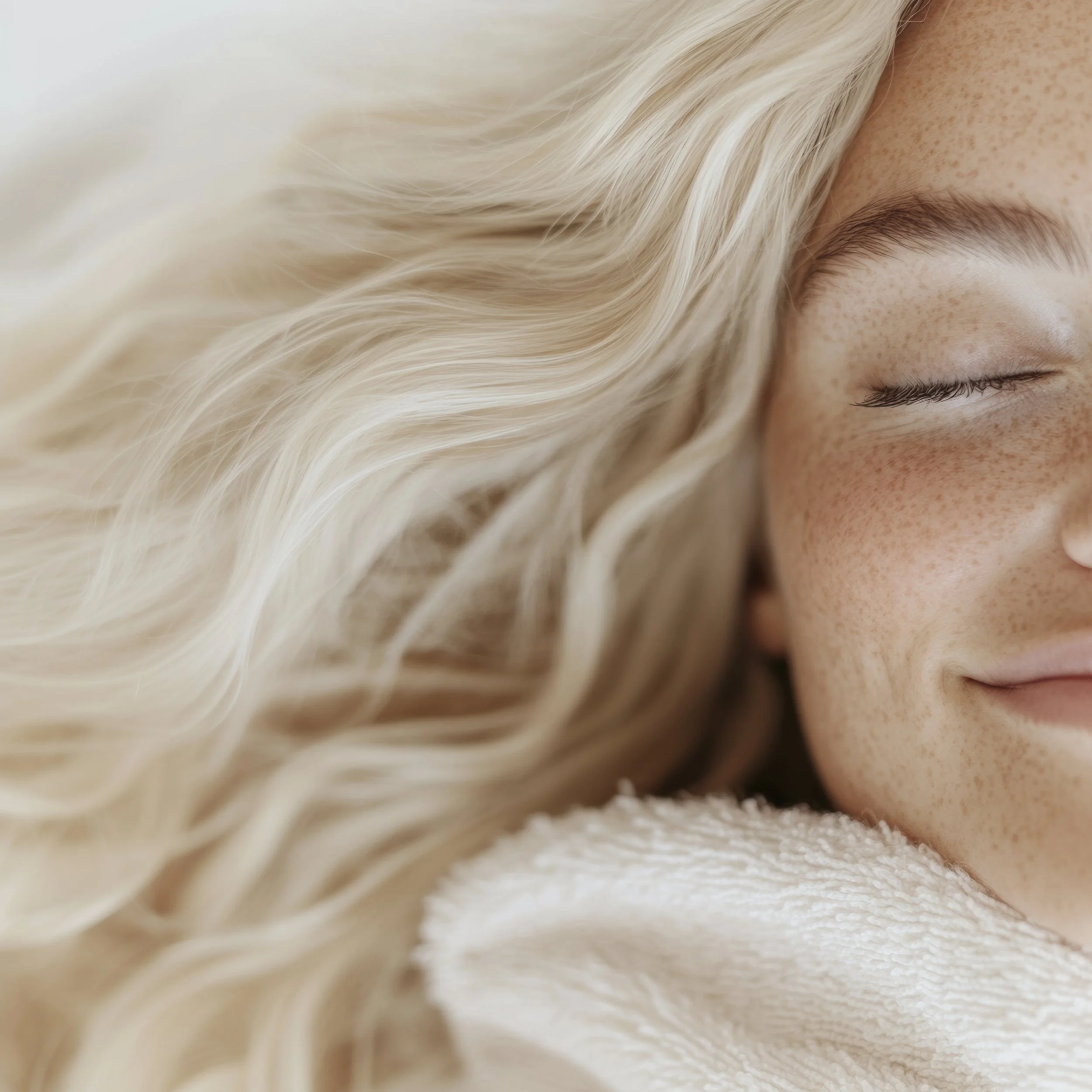 Close-up of a smiling woman with blonde, wavy hair, freckles, and closed eyes, resting her face on a soft, white surface.