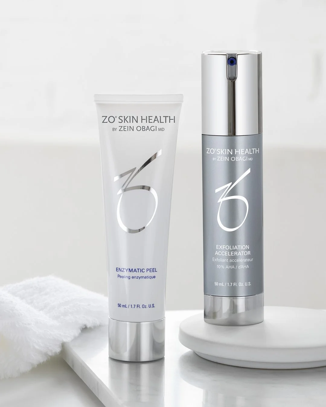 Two skincare products from ZO Skin Health by Zein Obagi MD displayed on a white surface. The left product is an enzymatic peel in a white tube, and the right is an exfoliation accelerator in a silver pump bottle. A white towel and a white round platform are also visible.