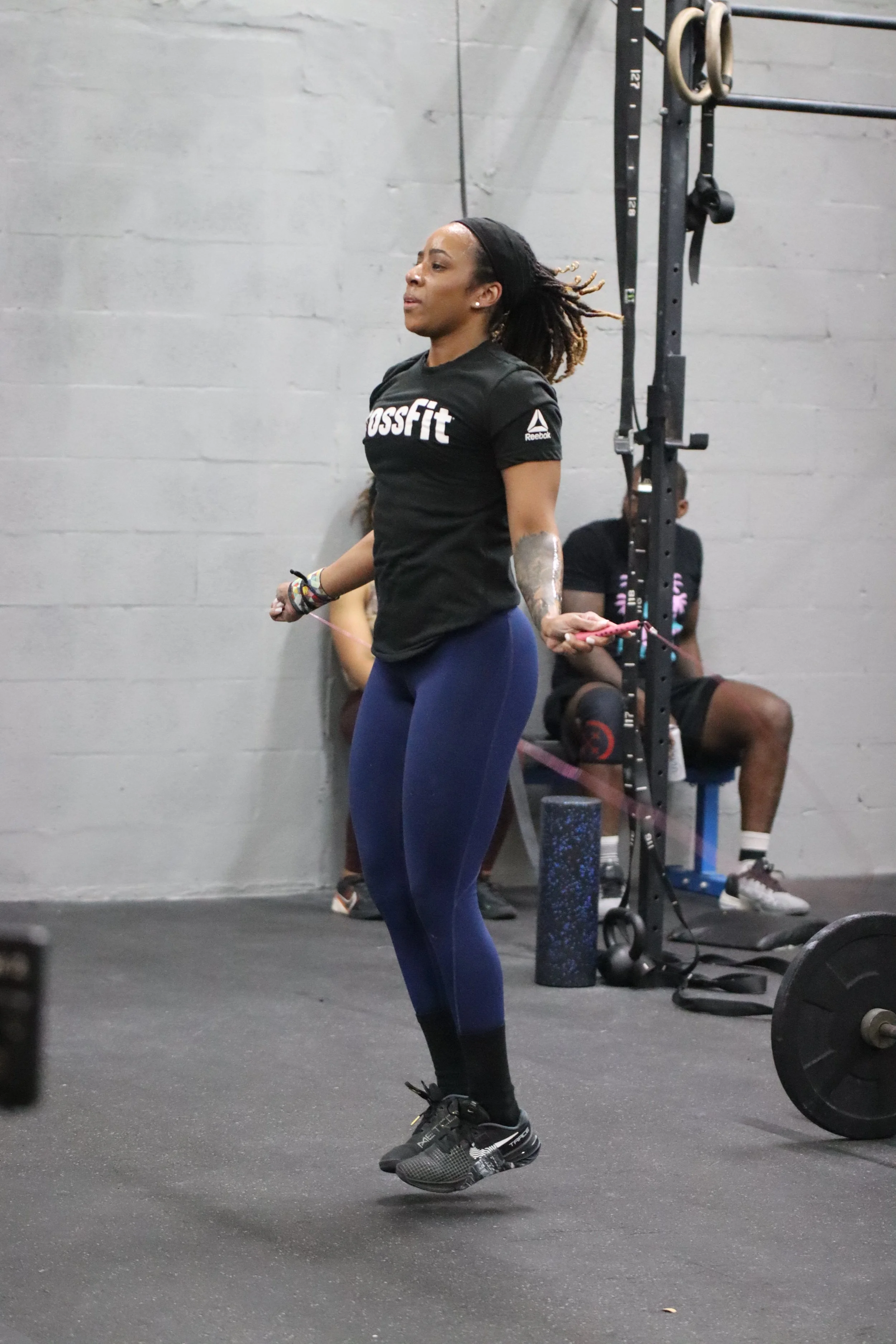 A woman with long dreadlocks jumping rope in a gym, wearing a black CrossFit shirt, blue leggings, and black athletic shoes.