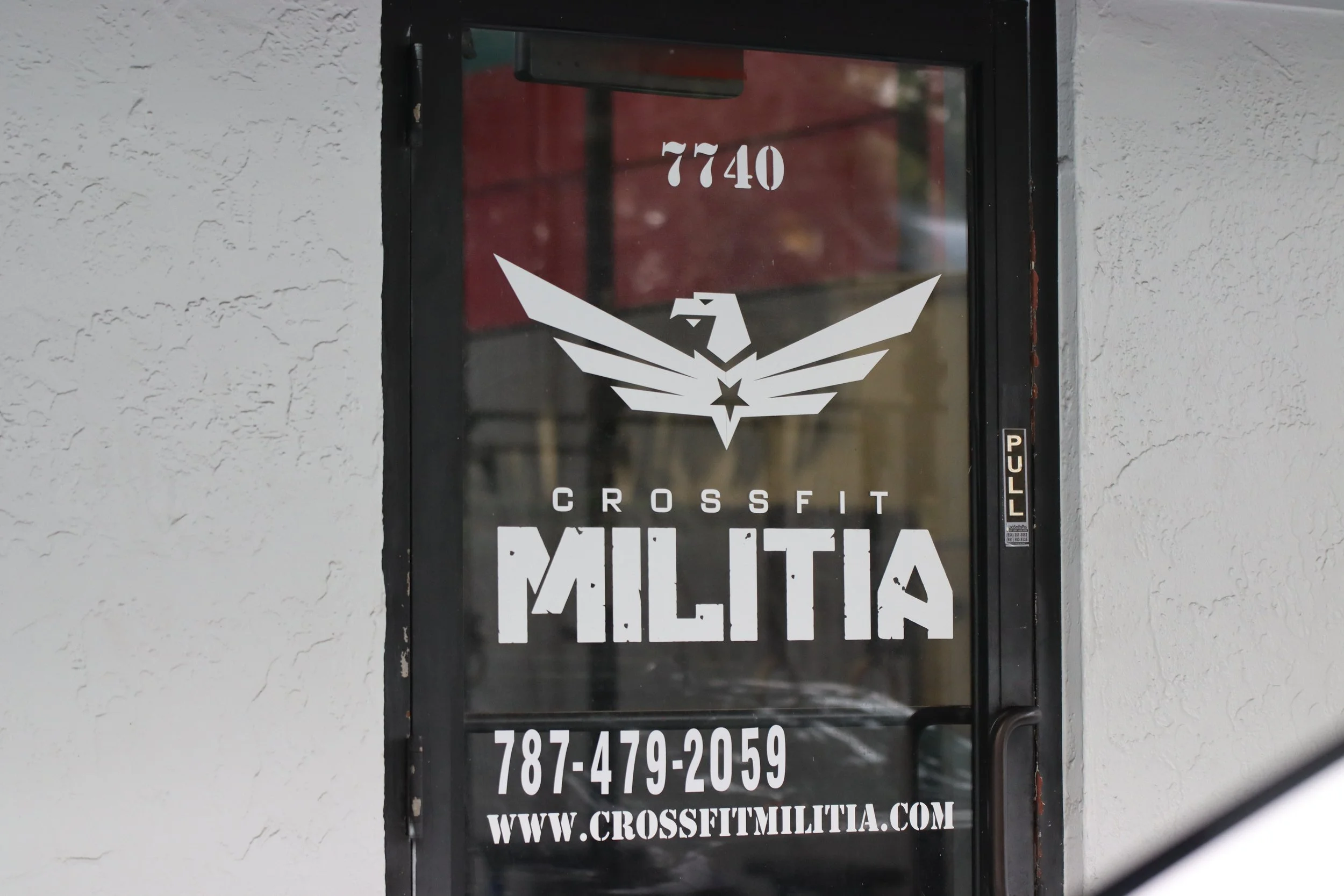 Glass door with the text 'CrossFit Militia,' a stylized eagle logo, phone number, website URL, and address number, all in white